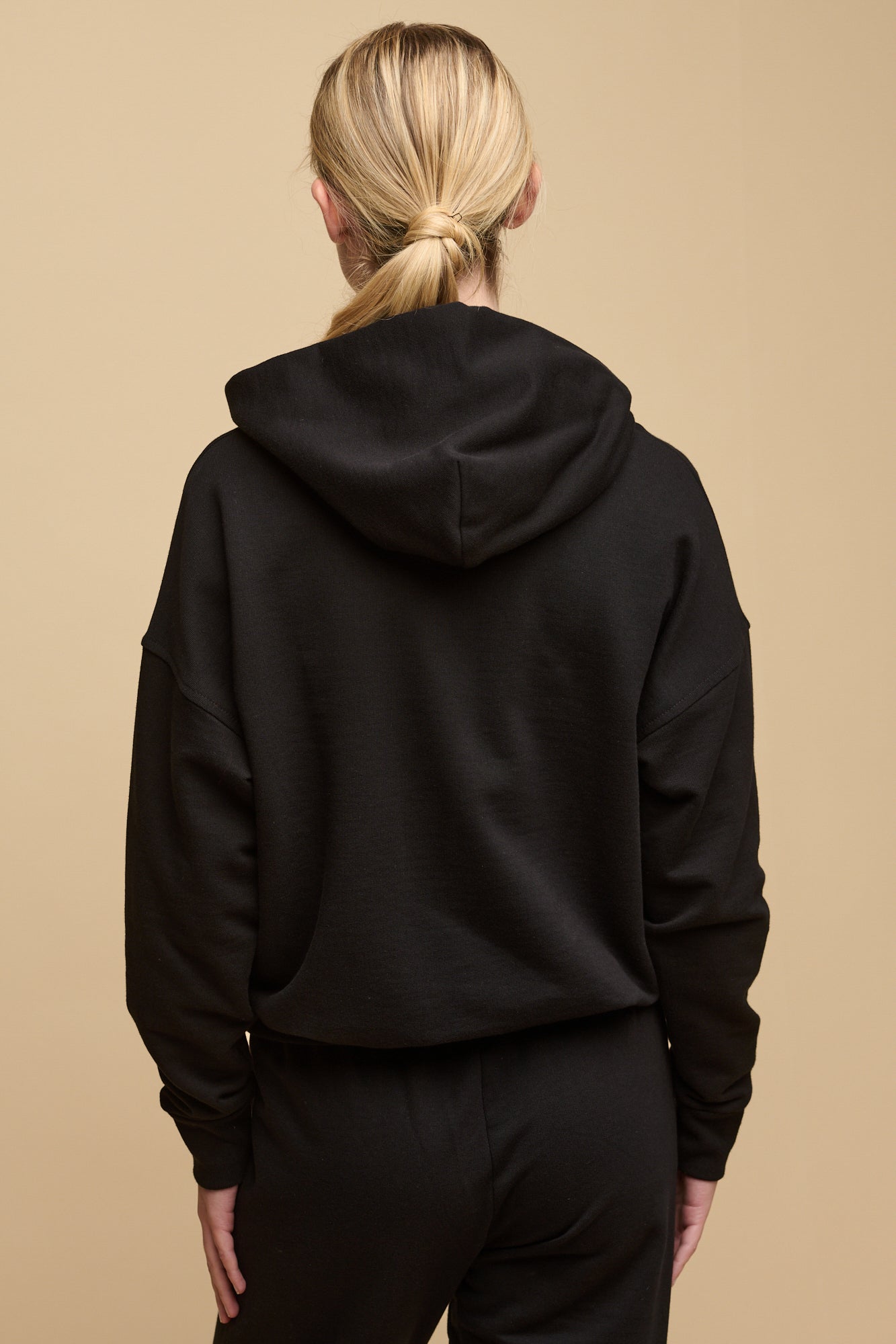 Female with blonde hair tied back wearing hooded sweatshirt in black with hands in front pocket. Ecru drawstring through the hood, worn over crew neck t shirt in white