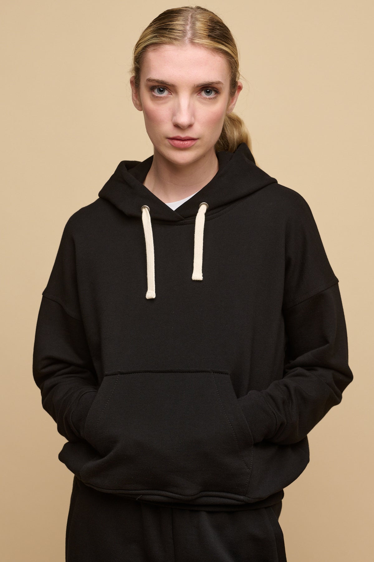 Female with blonde hair tied back wearing hooded sweatshirt in black with hands in front pocket. Ecru drawstring through the hood, worn over crew neck t shirt in white