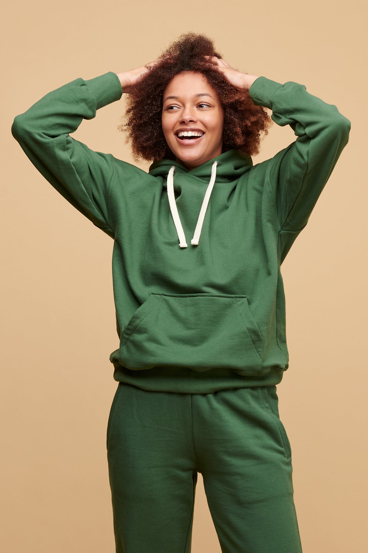 Thigh up of black female with an afro with her hands in her hair wearing 100% cotton hooded sweatshirt in bottle green paired with matching sweatpants