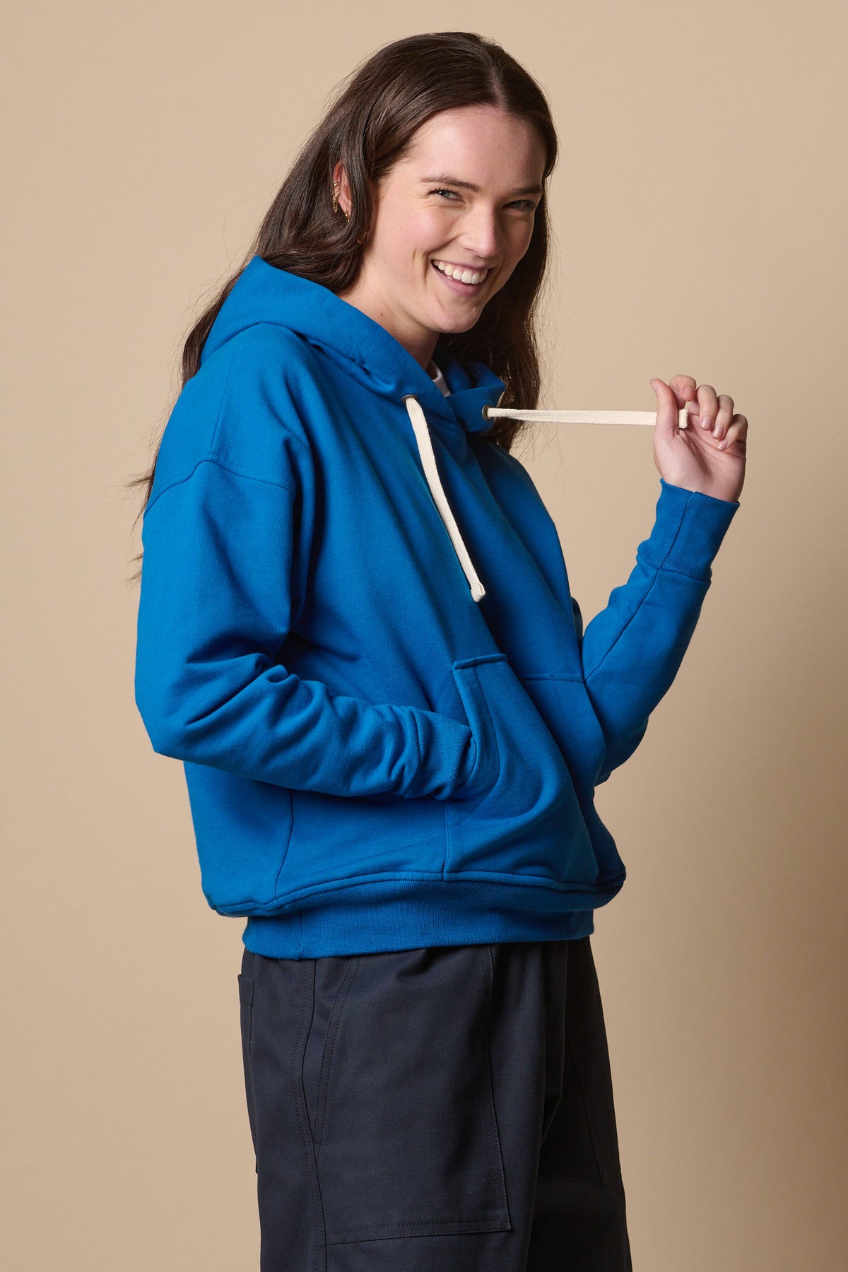 
            Smiley female wearing hooded sweatshirt in cobalt holding drawstring of the hood with one hand and one hand in the front pocket 