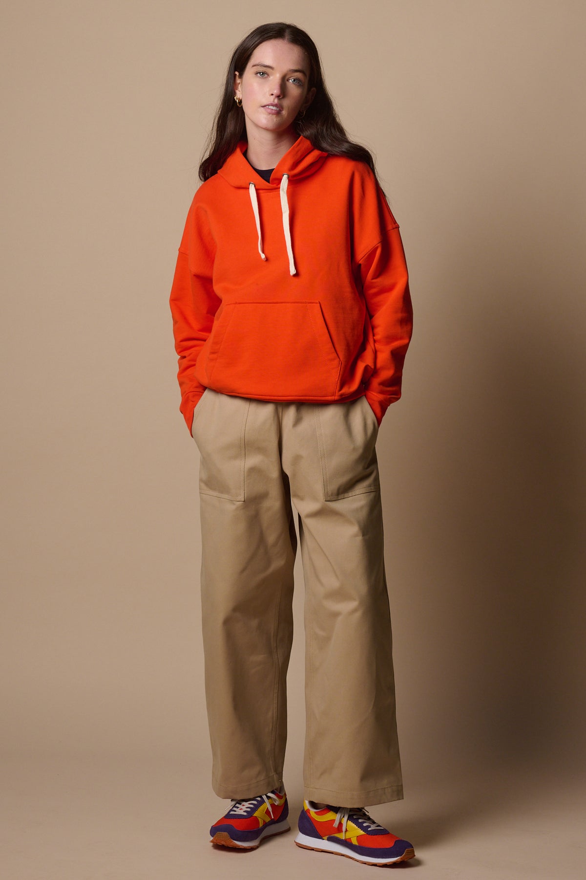 Full body image of white female wearing hooded sweatshirt in flame red, paired with camerawoman trousers in putty