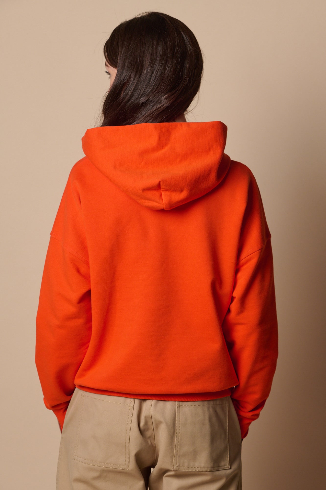 Full body image of white female wearing hooded sweatshirt in flame red, paired with camerawoman trousers in putty