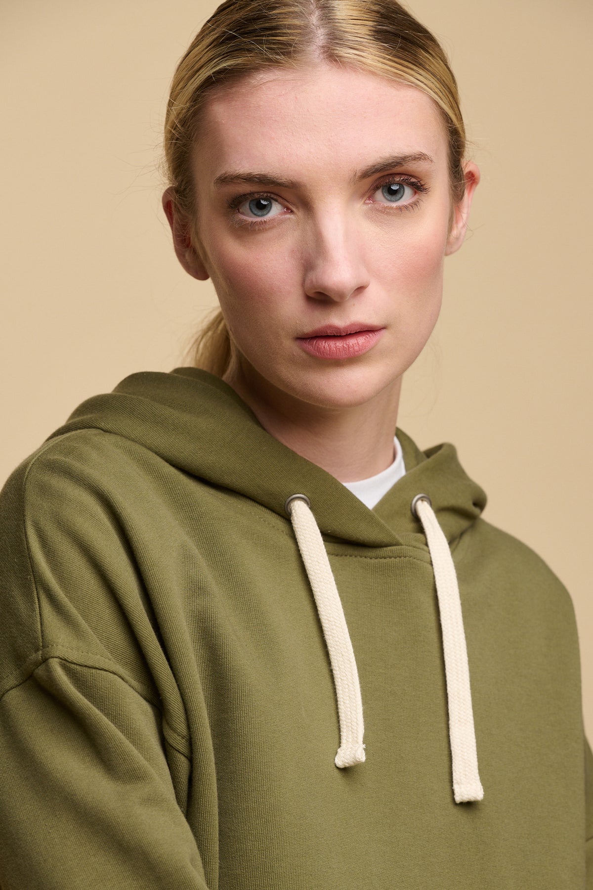 Portrait of blonde female wearing hooded sweatshirt in olive with ecru drawstring through the hood, worn over a crew neck t shirt in white