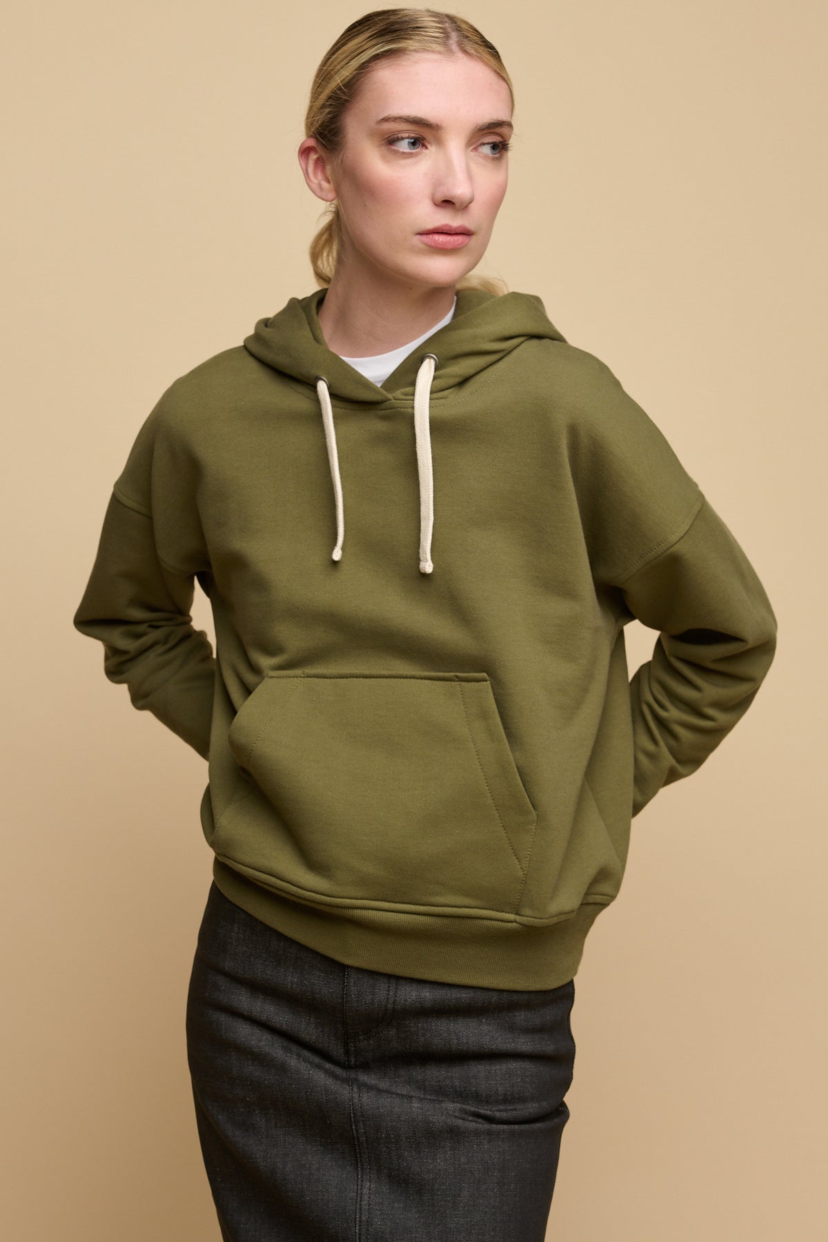 Thigh up image of blonde female wearing hooded sweatshirt in olive with one front pocket, worn over white crew neck t shirt paired with denim maxi skirt in black