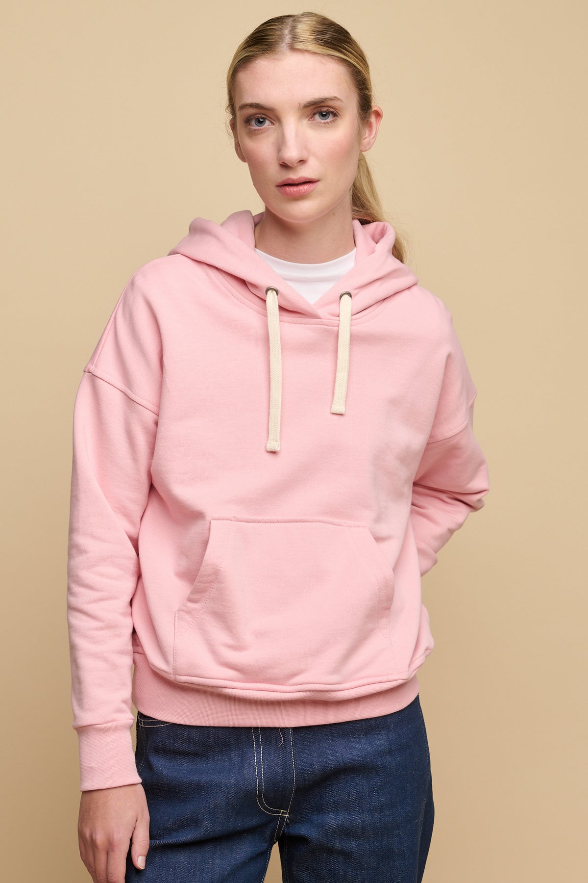 Thigh up image of the front of female wearing pale pink hooded sweatshirt paired with work jeans in blue