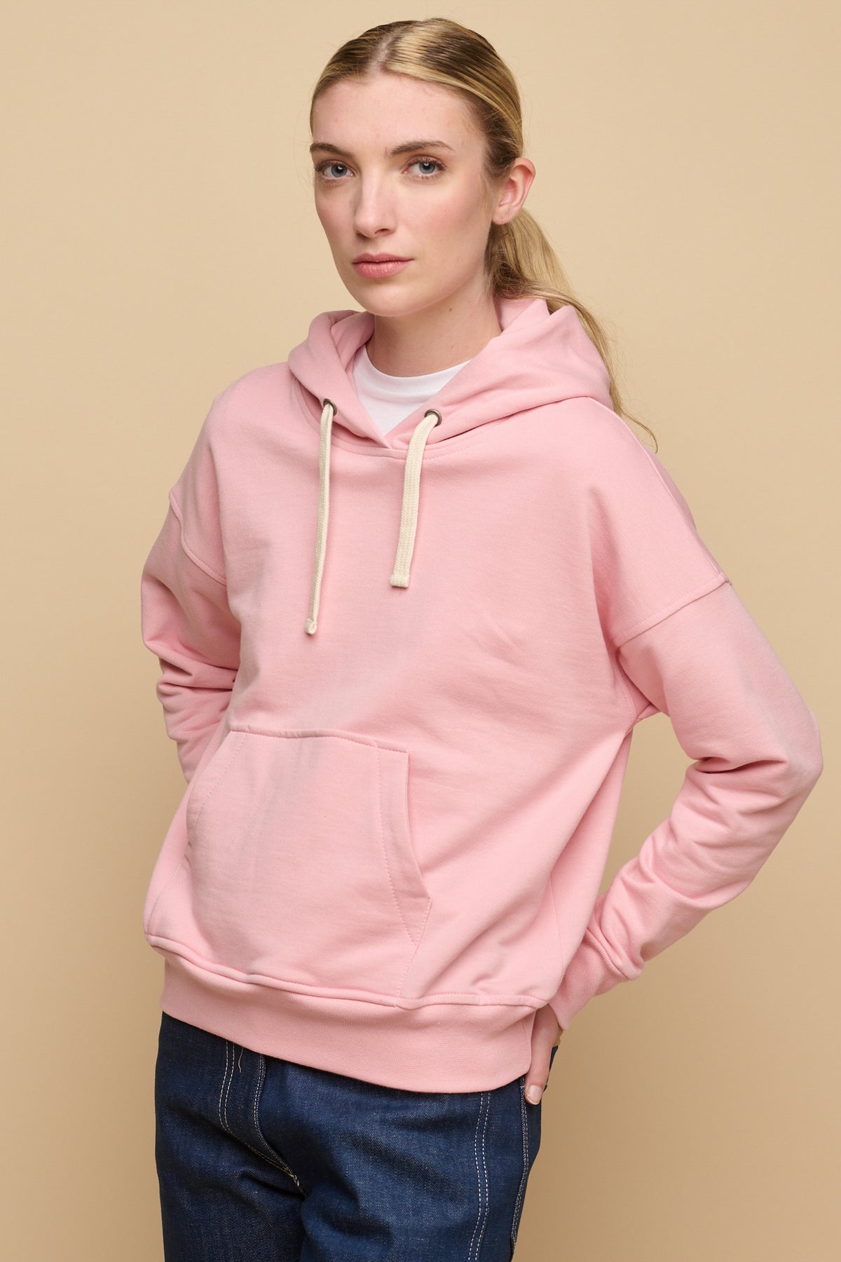 Thigh up image of the front of female wearing pale pink hooded sweatshirt paired with work jeans in blue