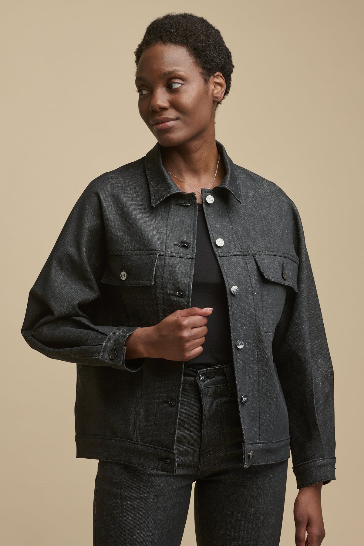 
            Thigh up front of female wearing Jodie denim jacket in black with two front pockets and two front chest pockets with falsp, button cuff and adjustable button fastening on the hem. Jacket unfastened over racer back vest in black