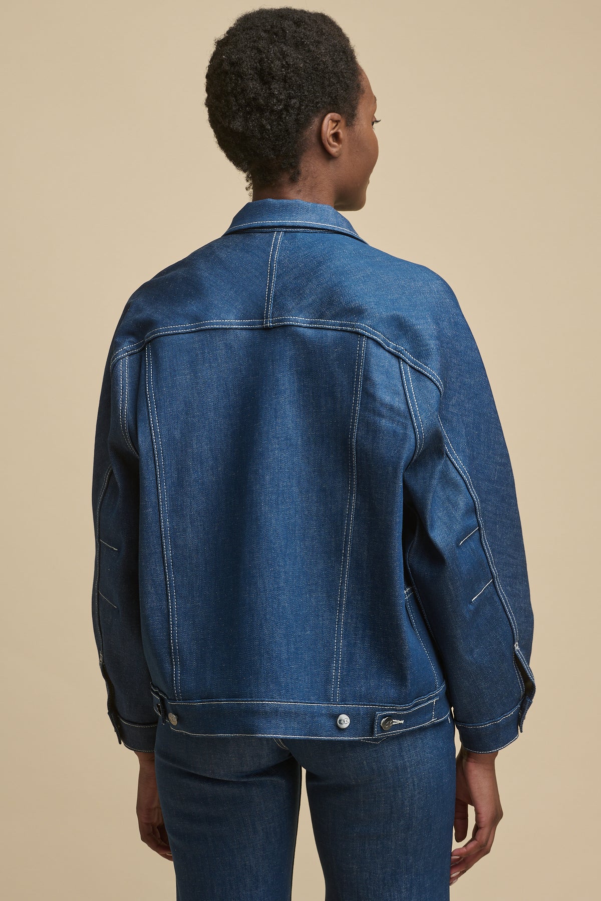 Thigh up image of the back of black female wearing Jodie denim jacket in blue with adjustable button fastening on hem. contrast stitching details