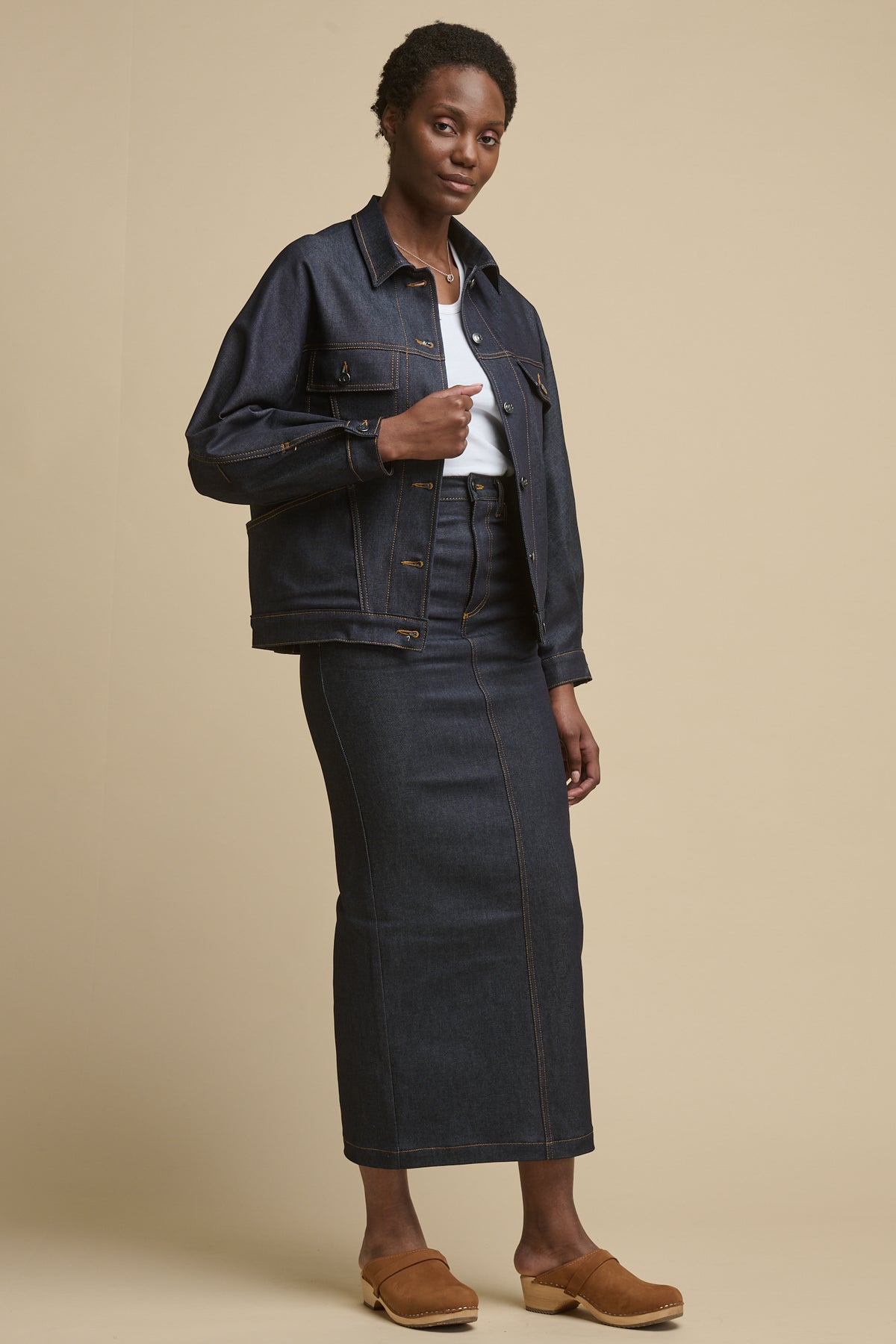 
            Full body image of female wearing Jodie denim jacket over white racer back vest paired with matching indigo denim Frankie maxi skirt