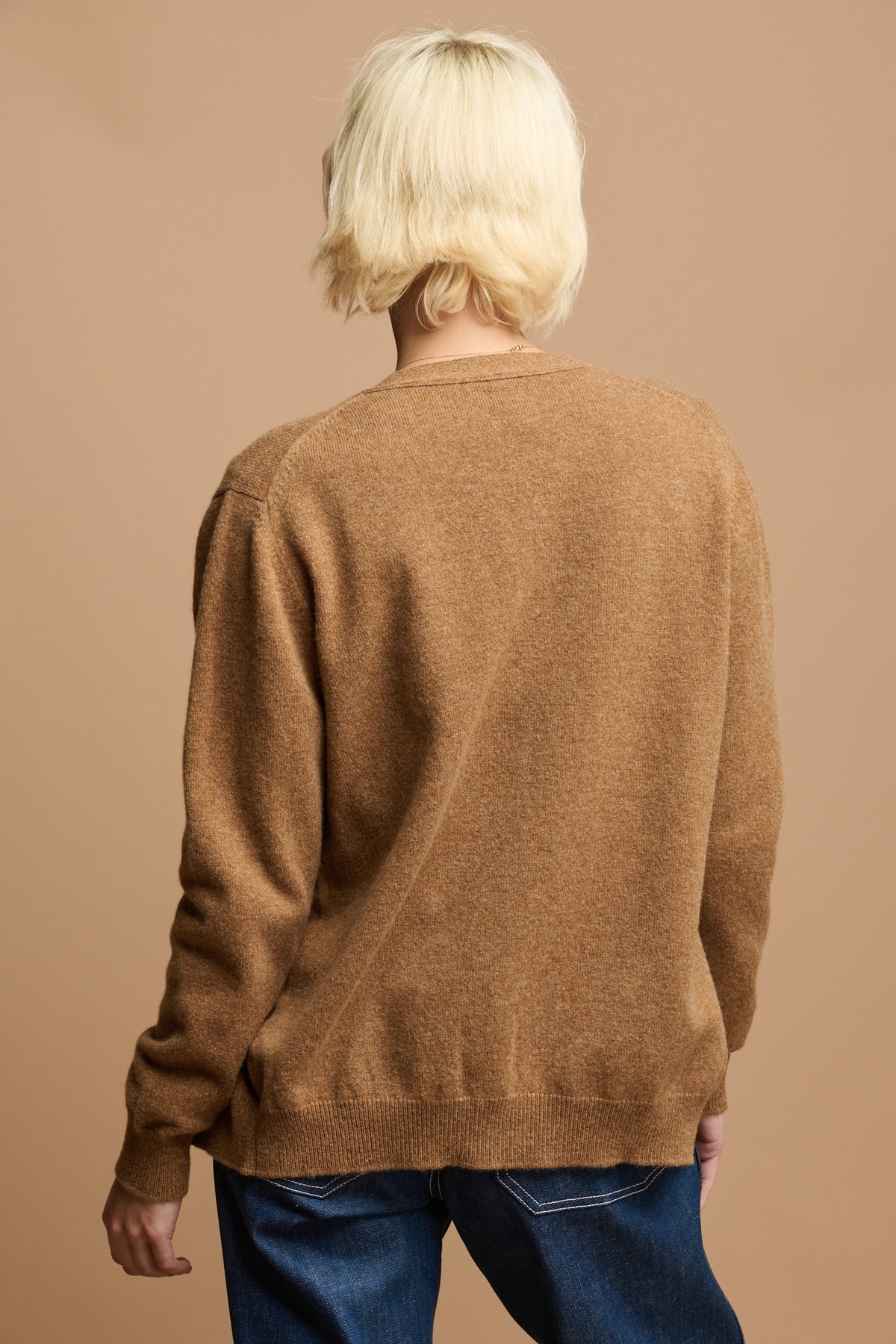 
            The back of female wearing lambswool cardigan in dark camel paired with work jeans in blue