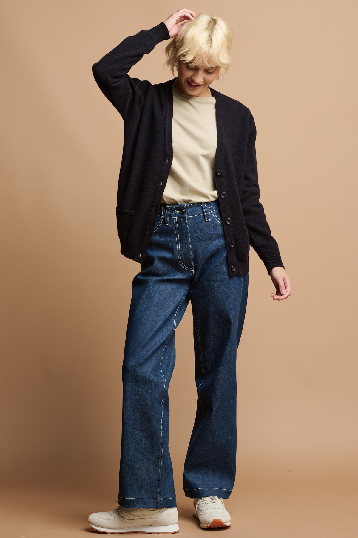 
            Full body front of female with one hand to her head wearing lambswool cardigan in navy unbuttoned over short sleeve t shirt in stone tucked into work jeans in blue