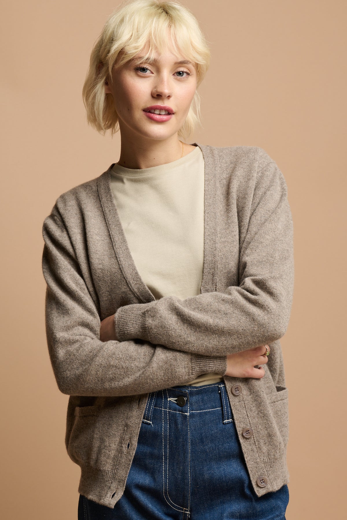 thigh up image of female with her arms folded wearing v neck lambswool cardigan in truffle unbuttoned over crew neck t shirt in stone