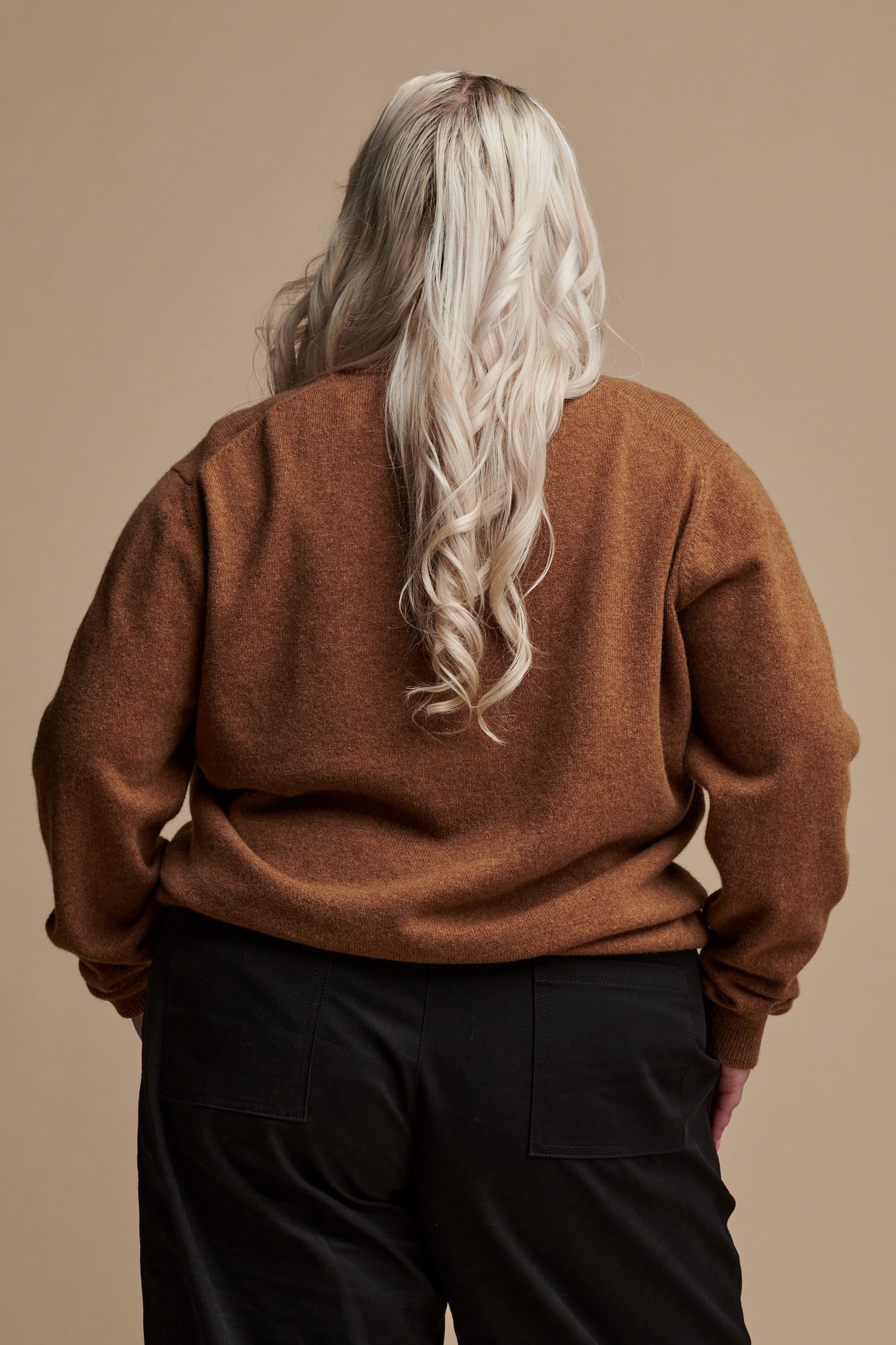 Thigh up of the back of female with long blonde hair wearing lambswool crew neck jumper in dark camel paired with camerawoman pants in black.