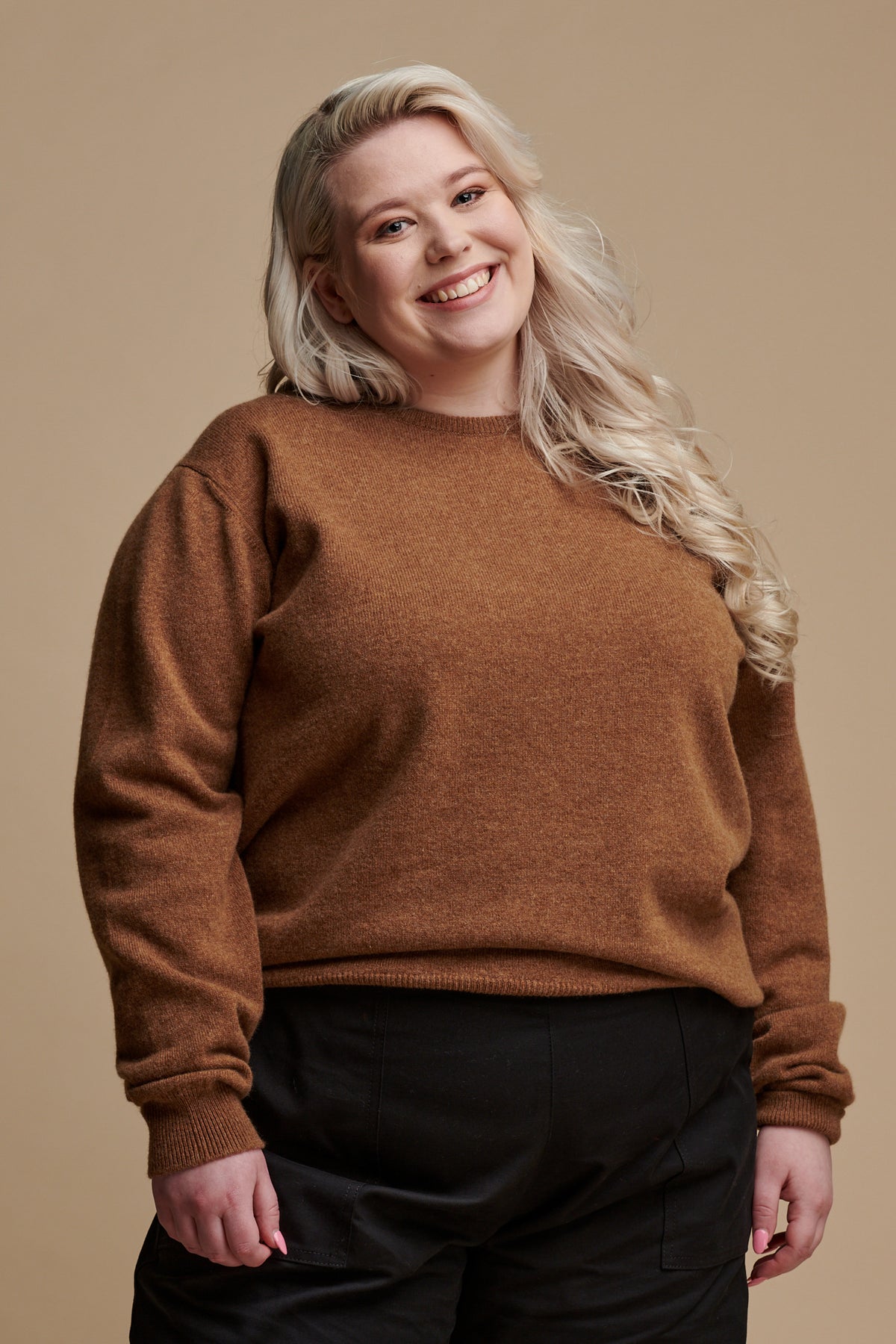 Thigh up of smiley female with long blonde hair wearing lambswool crew neck jumper in dark camel paired with camerawoman pants in black.