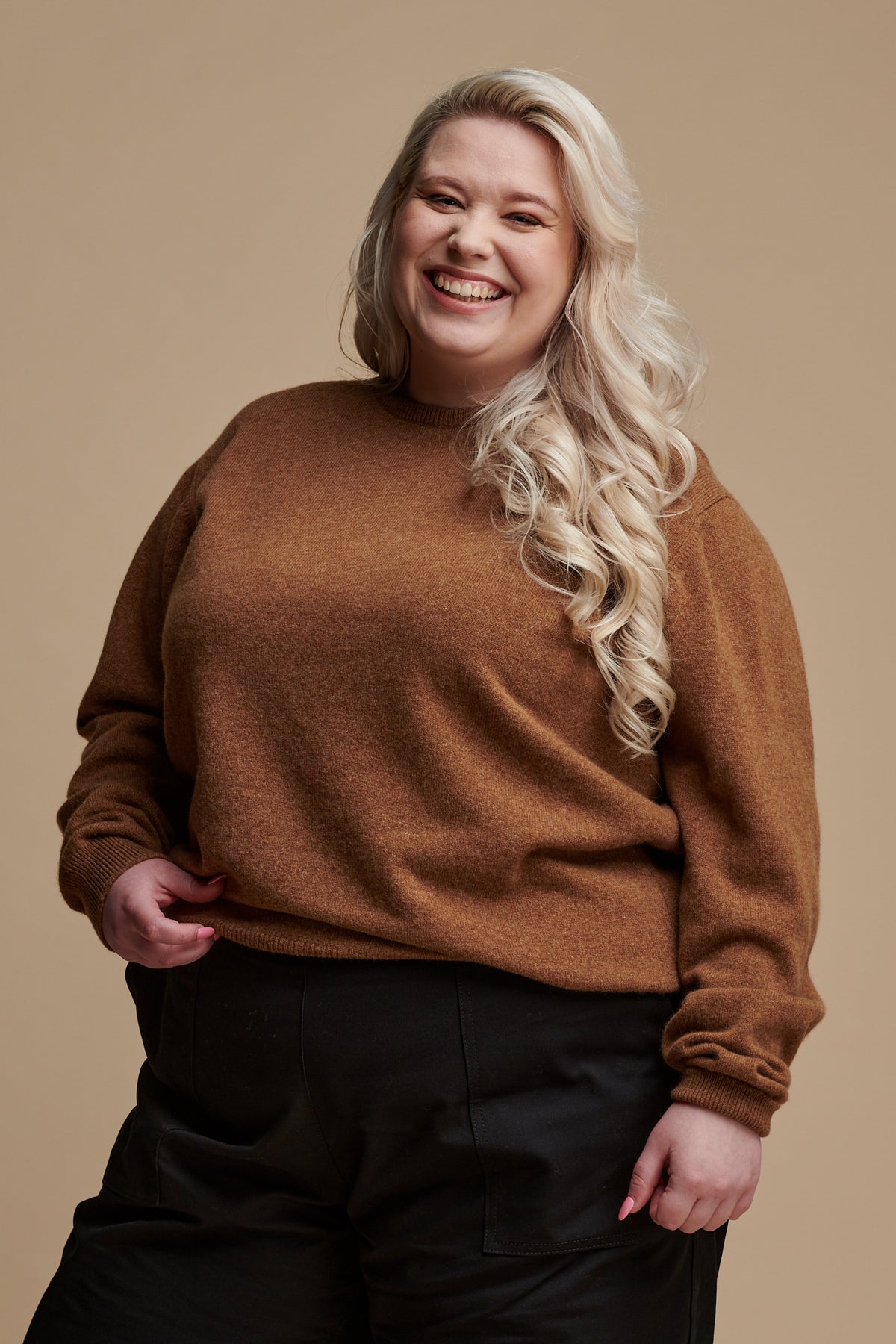 Thigh up of smiley female with long blonde hair wearing lambswool crew neck jumper in dark camel paired with camerawoman pants in black.