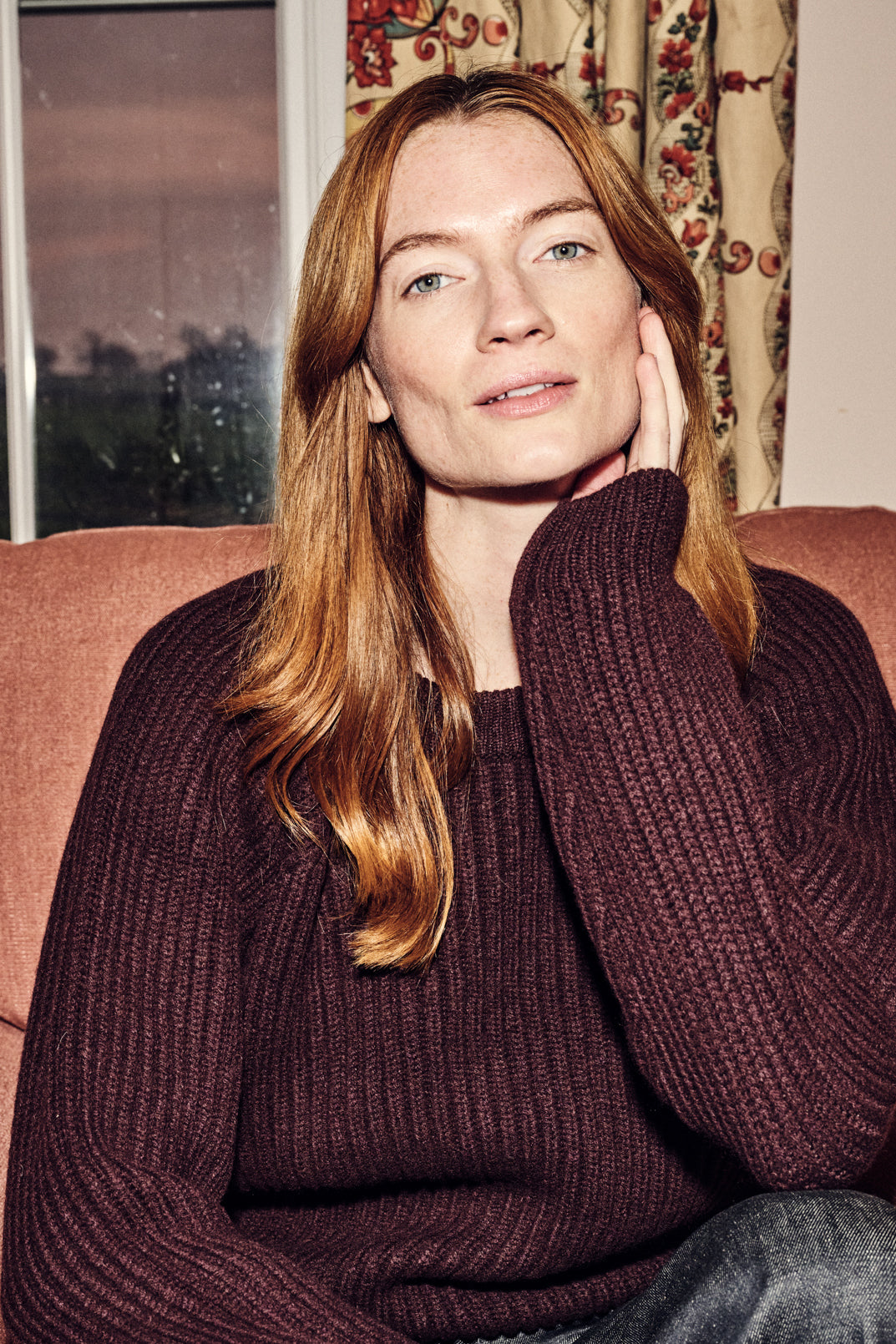 Woman wearing a brown jumper sitting on a couch with a window and curtains in the background.
