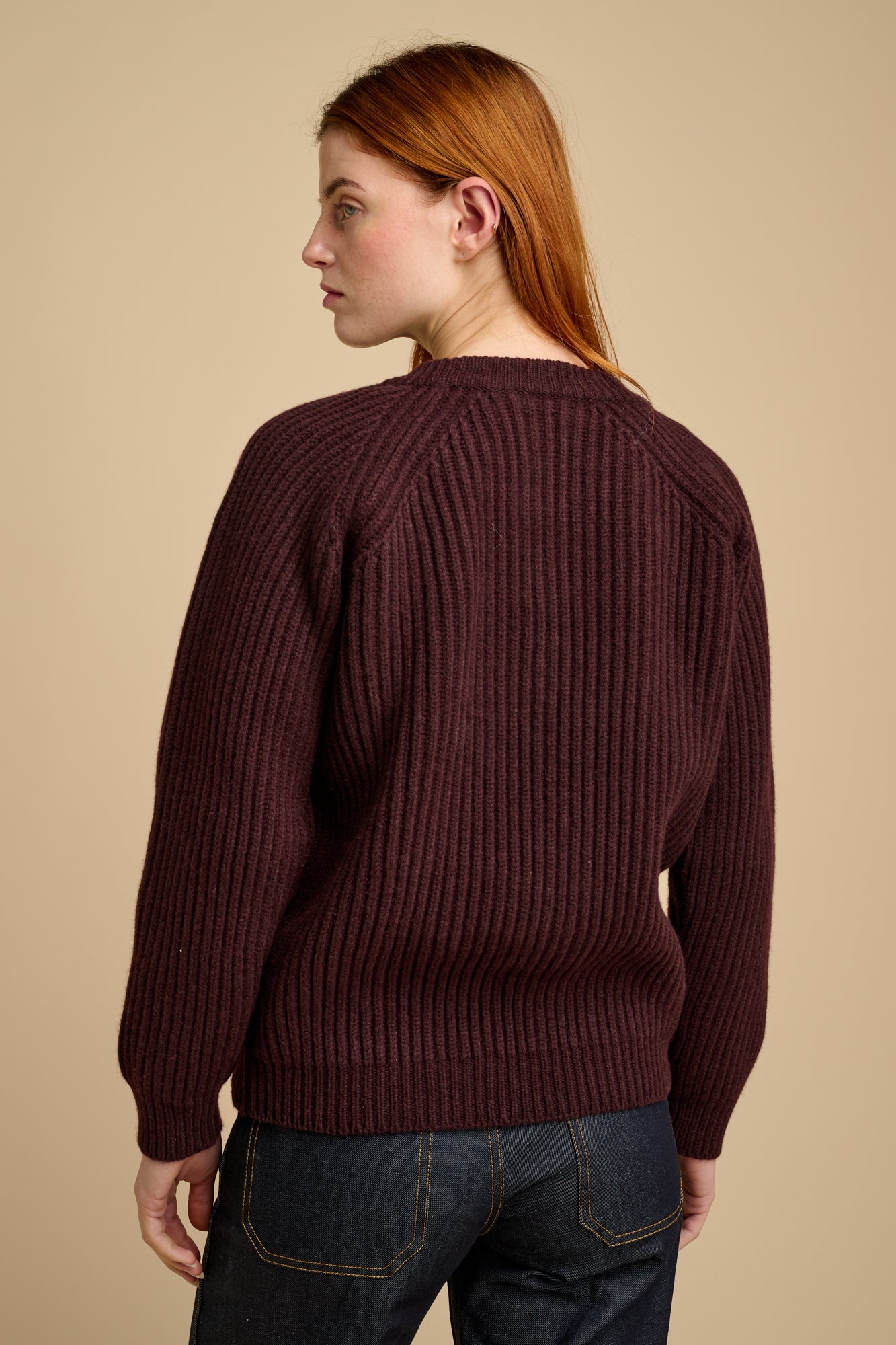 The back of woman wearing a lambswool fisherman ribbed jumper in chocolate against a beige background.