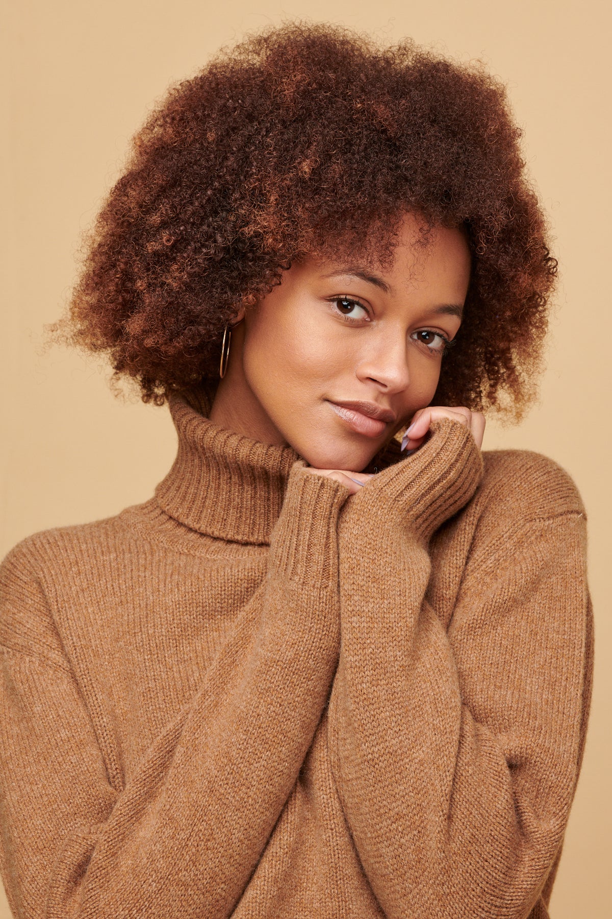 Portrait of female of colour with an afro wearing lambswool roll neck in dark camel holding her hands up under her chin