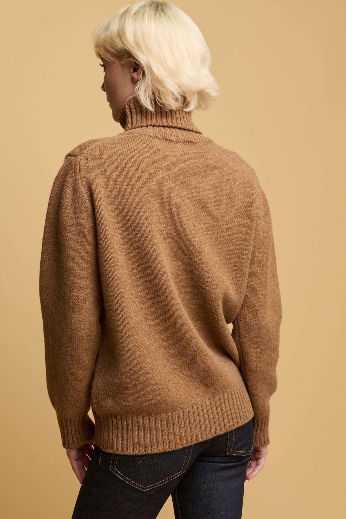 
            Thigh up of the back of female wearing lambswool roll neck in dark camel with ribbed hem