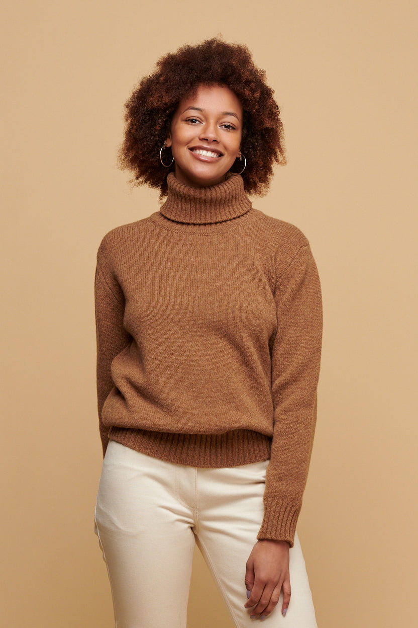 Thigh up of smiley black female with an afro wearing lambswool roll neck in dark camel paired with Community Clothing Gloria jeans in ecru