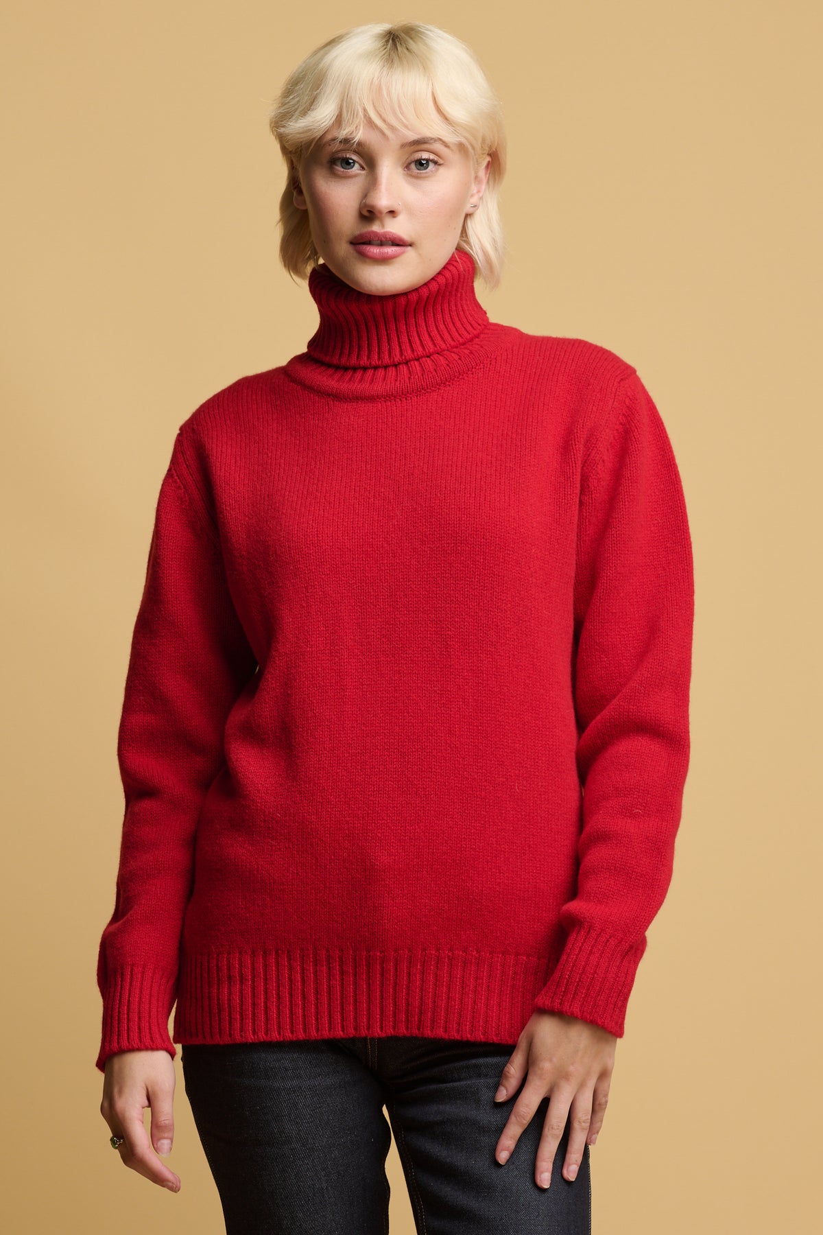 Thigh up of blonde female wearing lambswool roll neck in red with ribbed neck, cuff and hem paired with indigo jeans