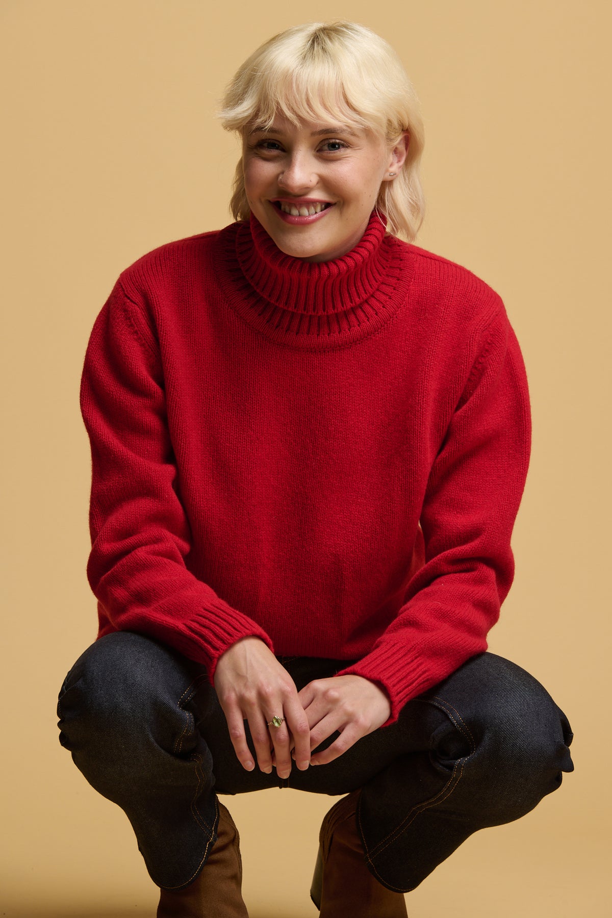 
            Smiley blonde female crouching down with her arms resting on her thighs wearing lambswool roll neck jumper in red