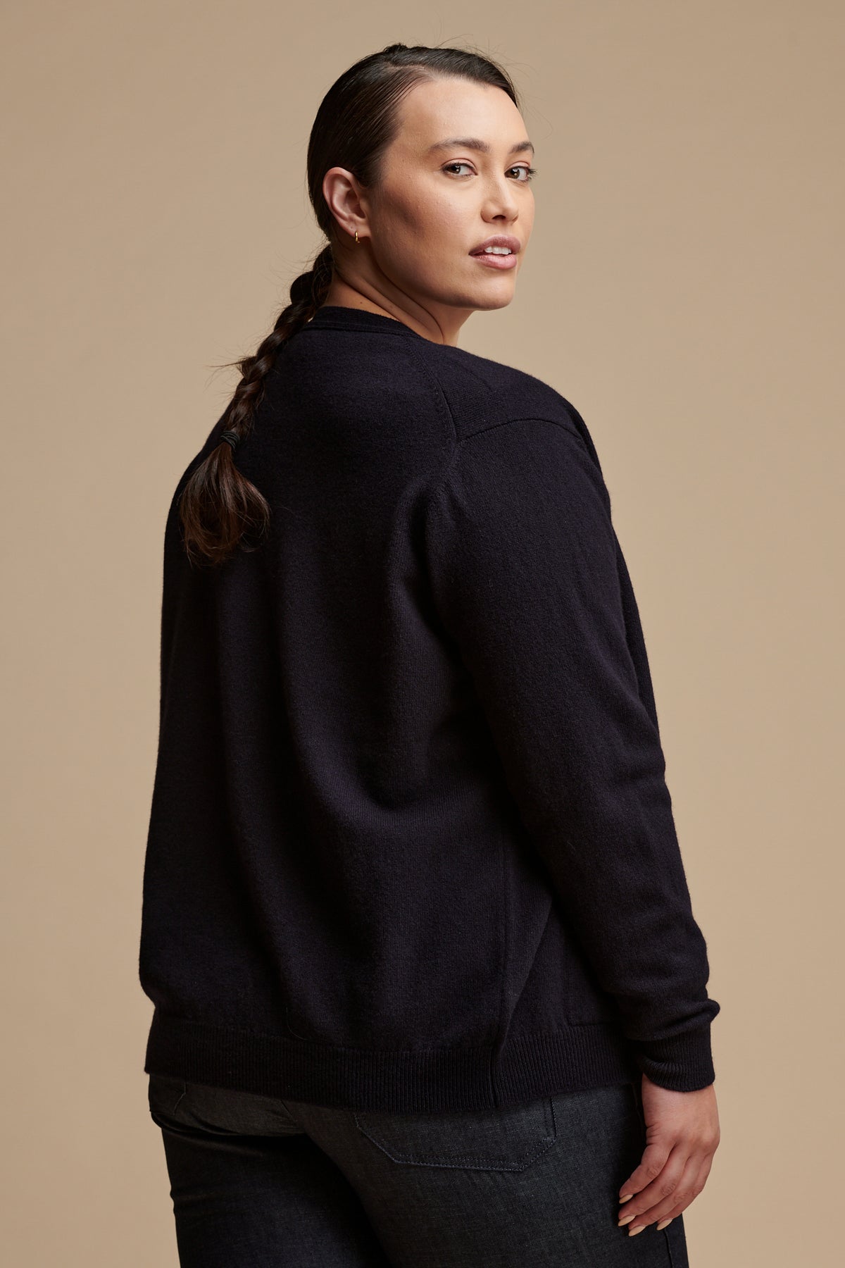 Thigh up image of the back of female with her hair tied in a low plait wearing lambswool v neck cardigan in navy paired with work trousers in indigo.