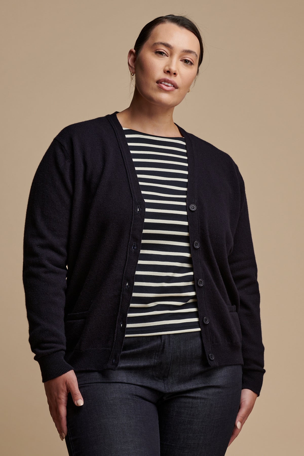 Thigh up image of female wearing lambswool v neck cardigan in navy unbuttoned over Breton top in navy-ecru, paired with work trousers in indigo.
