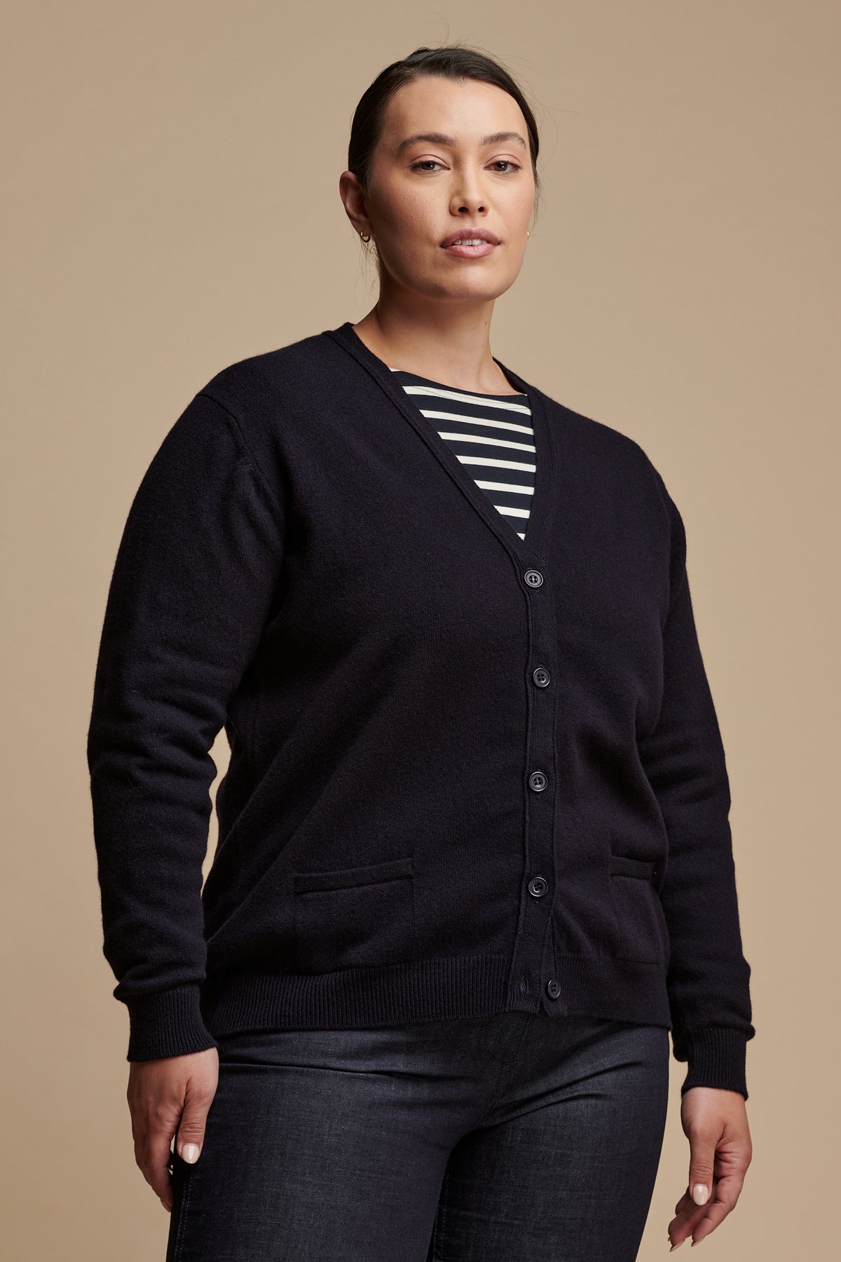 Thigh up image of female wearing lambswool v neck cardigan in navy buttoned up over Breton top in navy-ecru, paired with work trousers in indigo.