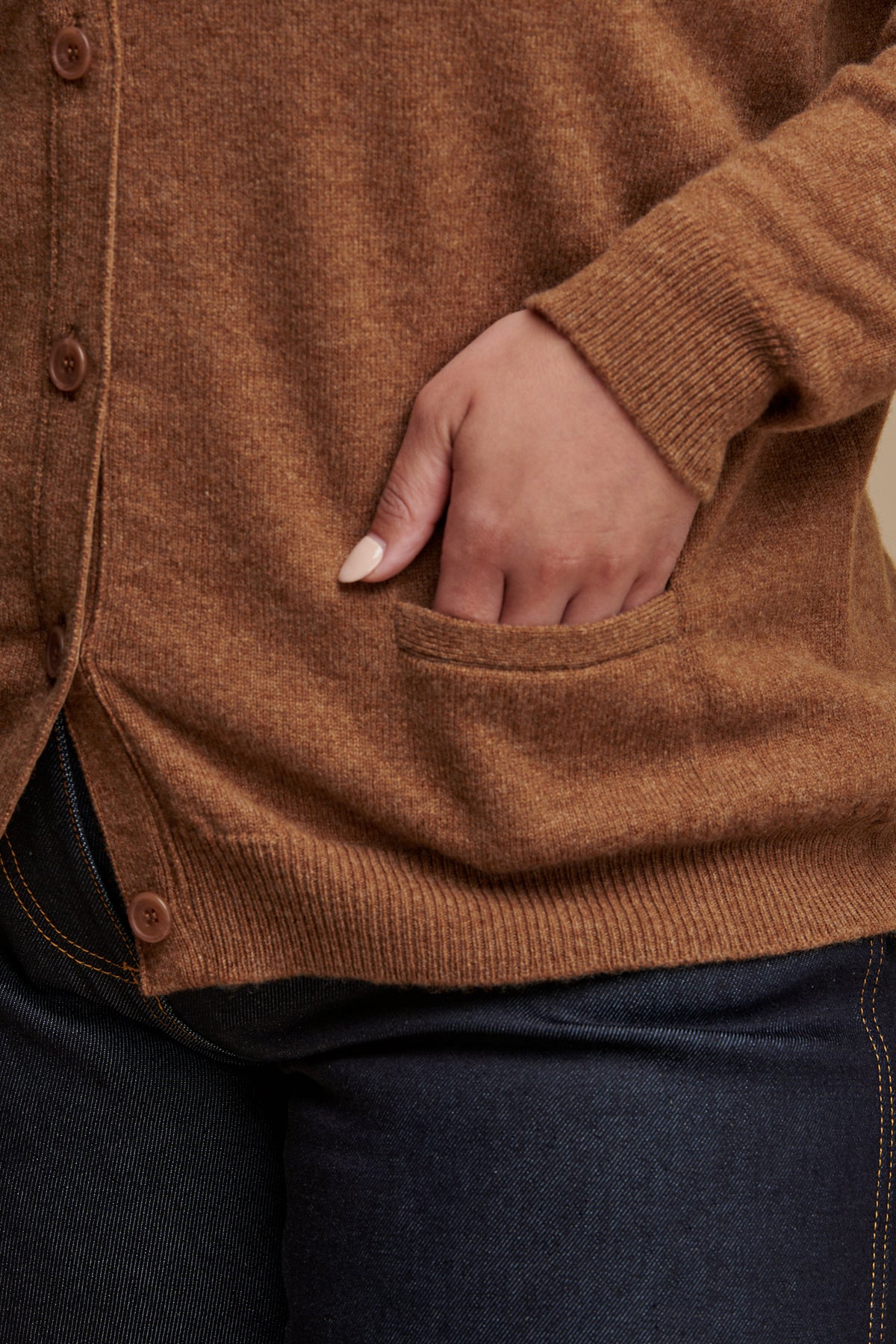 
            Close up of females hand in the pocket of lambswool v neck cardigan in dark camel, rib cuff and hem details.