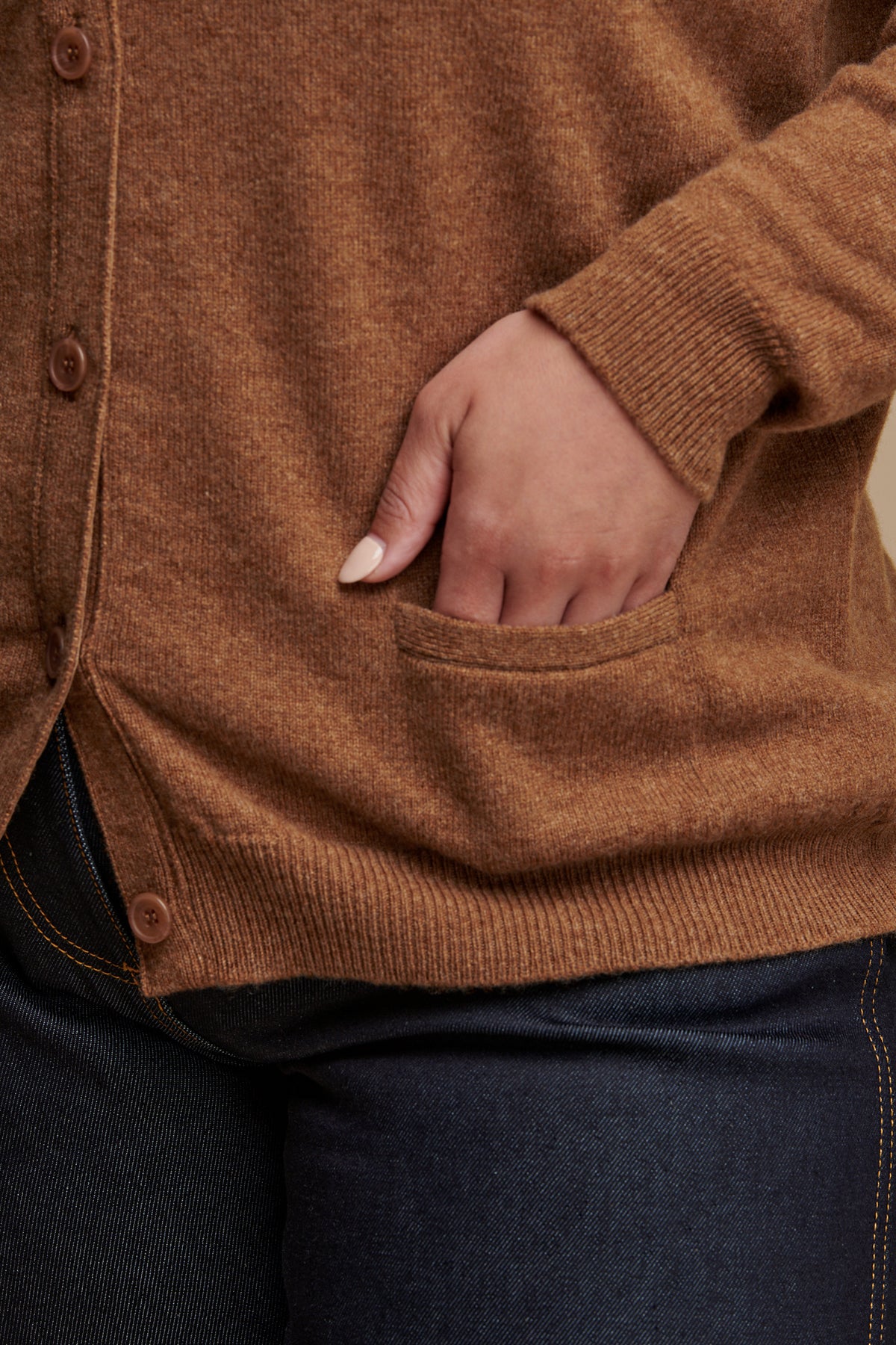 Close up of females hand in the pocket of lambswool v neck cardigan in dark camel, rib cuff and hem details.