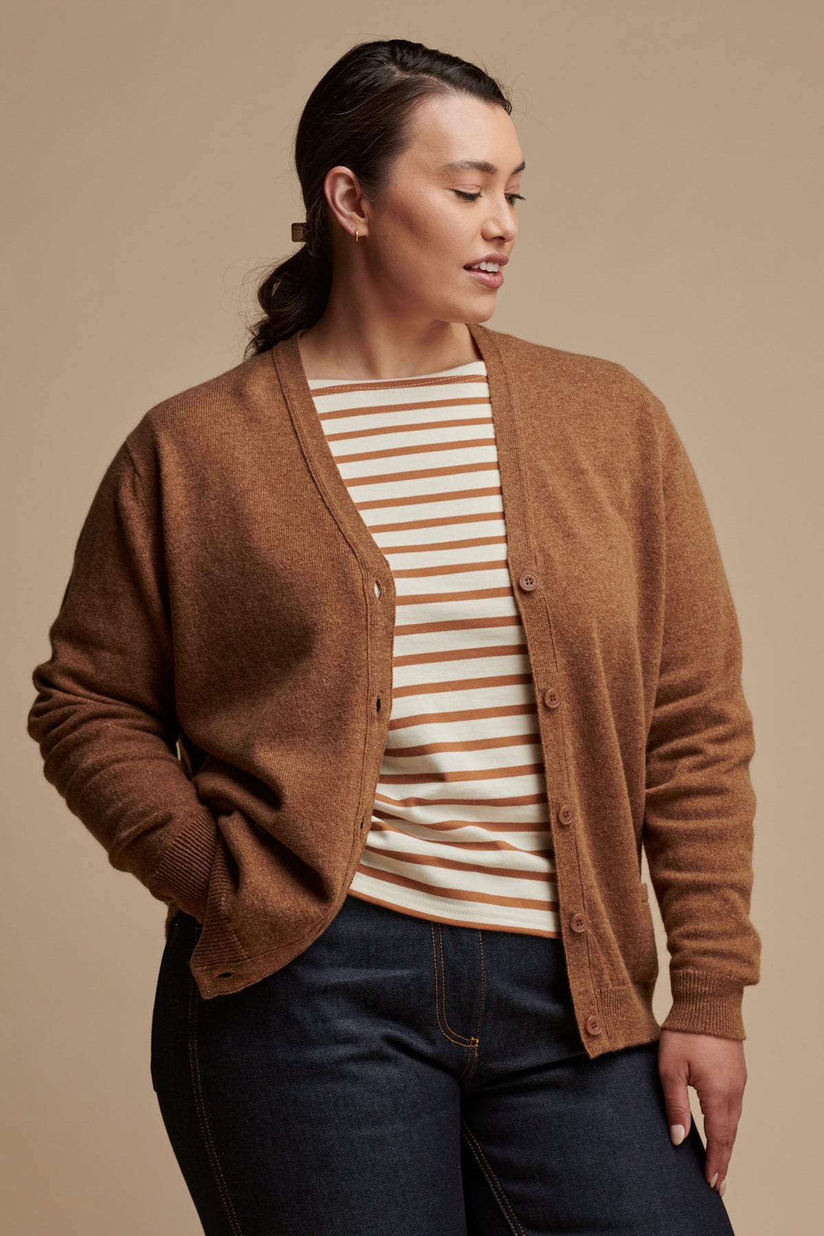 Thigh up image of female with her hand in the front jeans pocket wearing lambswool v neck cardigan in dark camel unbuttoned over Breton top in ecru-cinnamon, paired with indigo jeans.