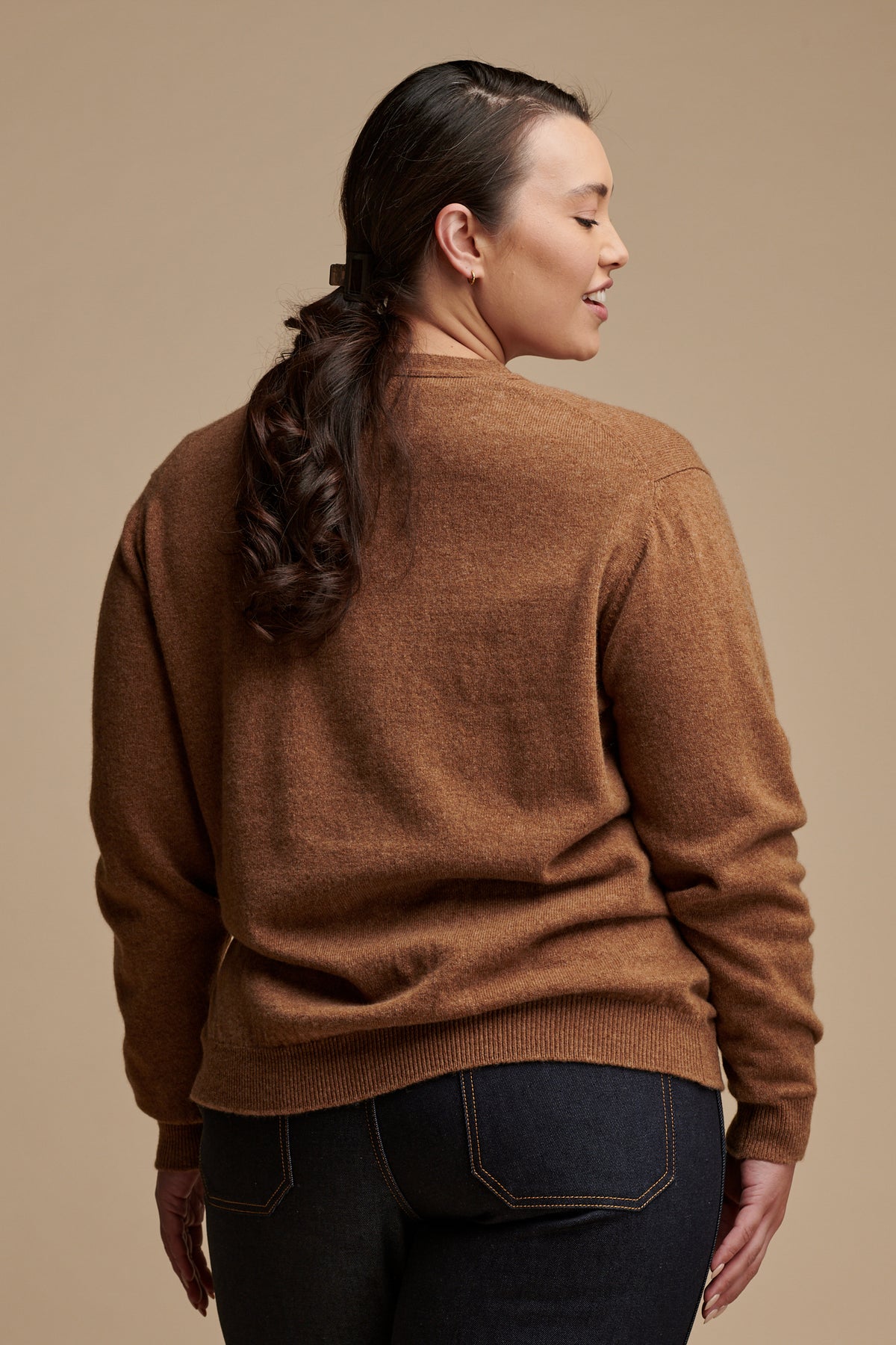 Thigh up image of the back of female with her her hair tied back into a low ponytail wearing lambswool v neck cardigan in dark camel, paired with indigo jeans.