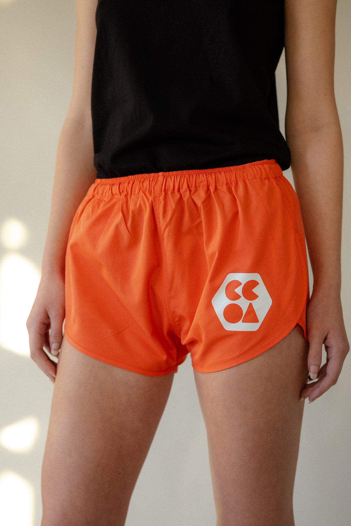 Image showing shoulder to knee of white female wearing lightweight running short plastic free in flame red, with white CCOA logo. Shorts have elasticated waist.