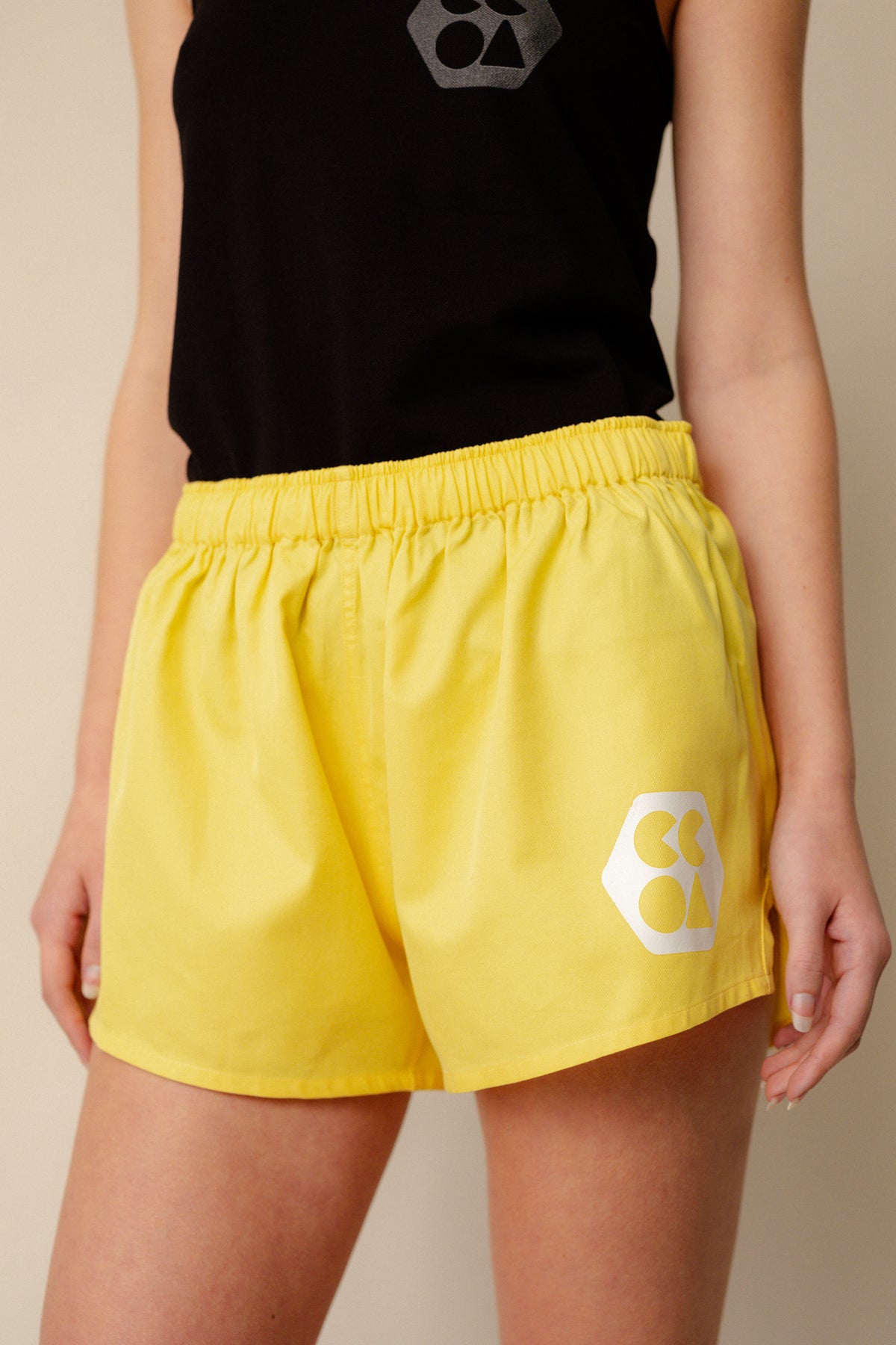 
            Shot of white, slim female from shoulder to thigh wearing lightweight sports short plastic free canary yellow with white CCOA logo