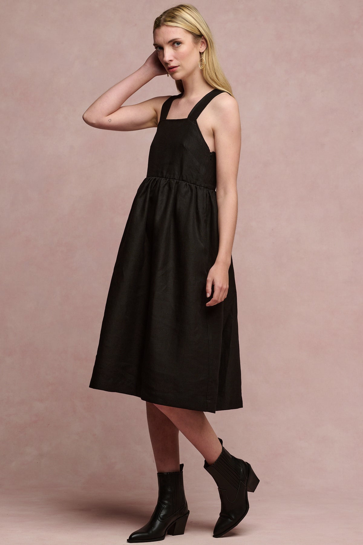 Full body image of fair skinned, blonde female wearing linen sun dress in black paired with black heeled boots
