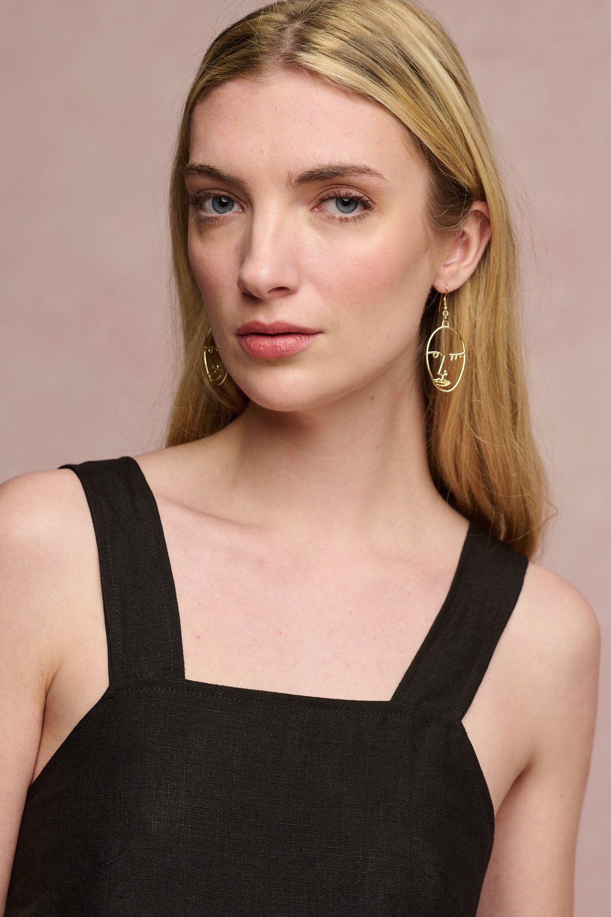 
            Portrait of fair skinned female with blonde hair behind ears wearing linen sun dress in black with gold earrings.