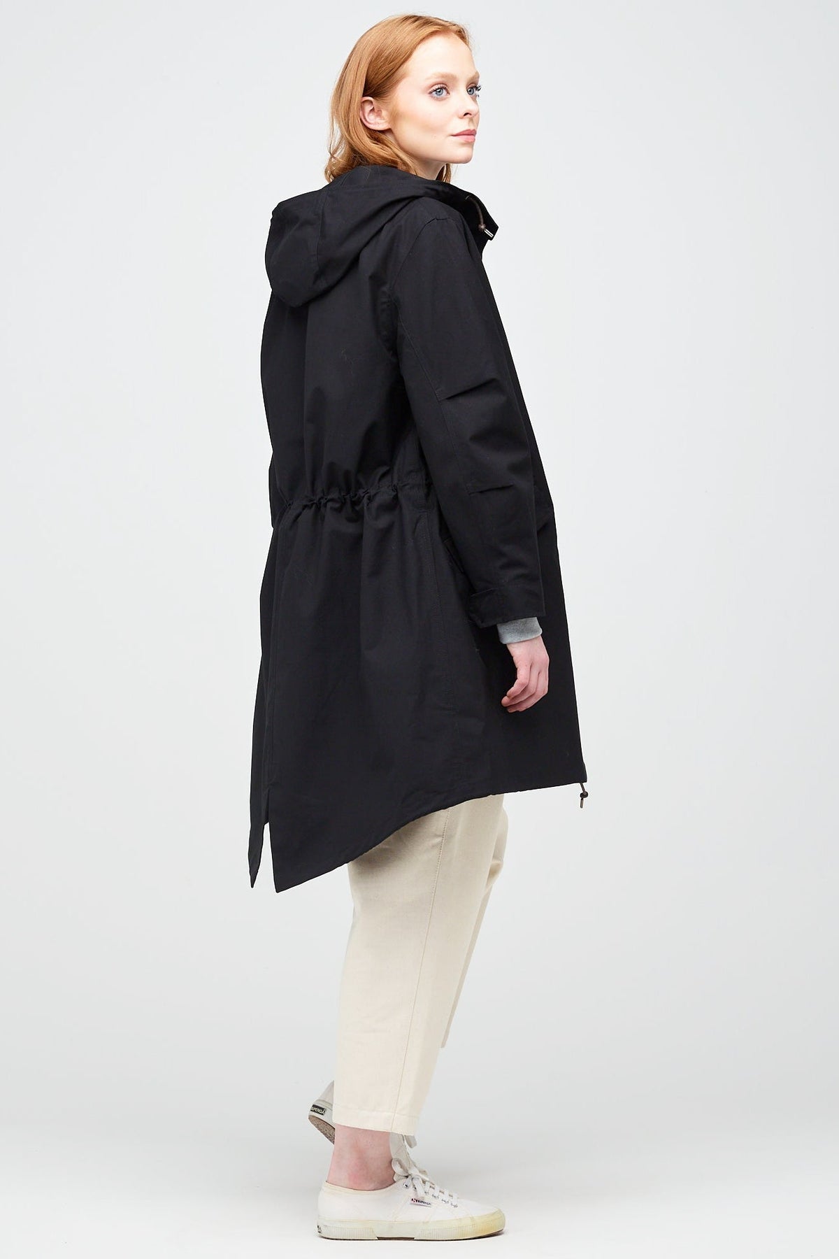 
            rear shot of a a young, white female model with ginger hair wearing a long parka in black. Styled with ecru jeans and a white trainer.