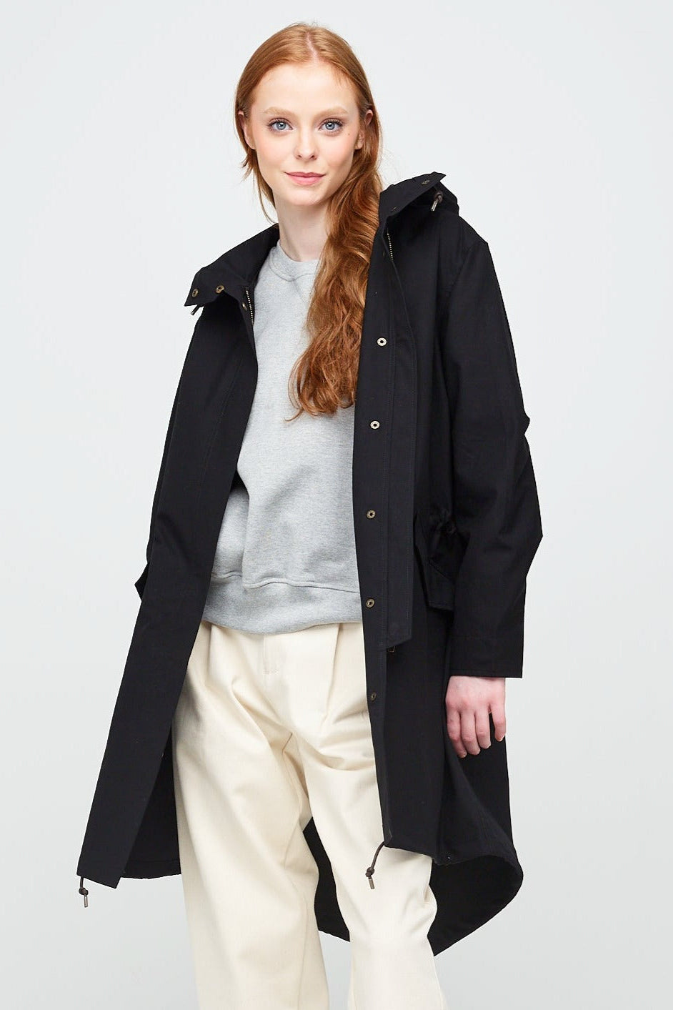 
            A young, white female model with ginger hair wearing long black parka unzipped. The parka is styled with a grey sweatshirt, ecru jeans and a white trainer.