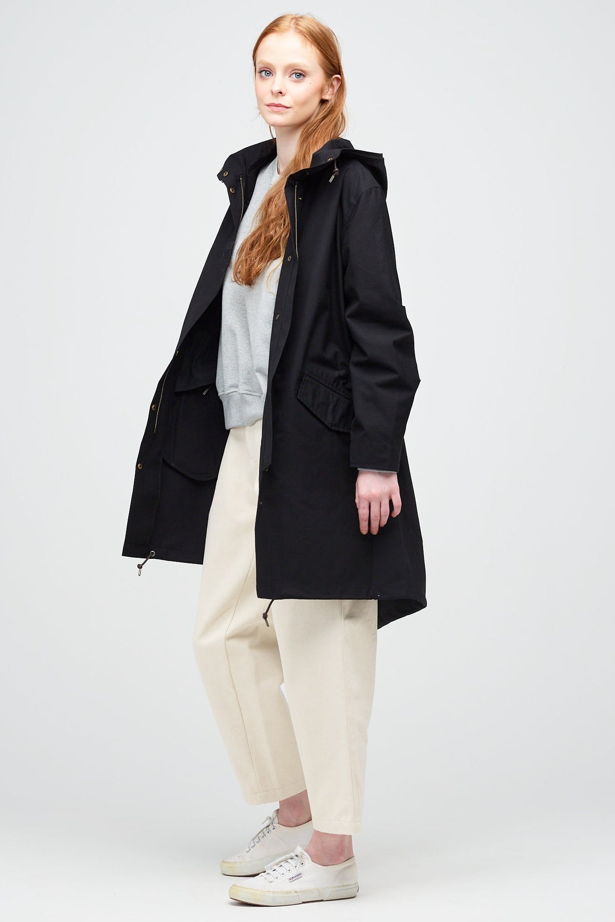 
            Side shot of A young, white female model with ginger hair wearing long black parka unzipped. The parka is styled with a grey sweatshirt, ecru jeans and a white trainer.