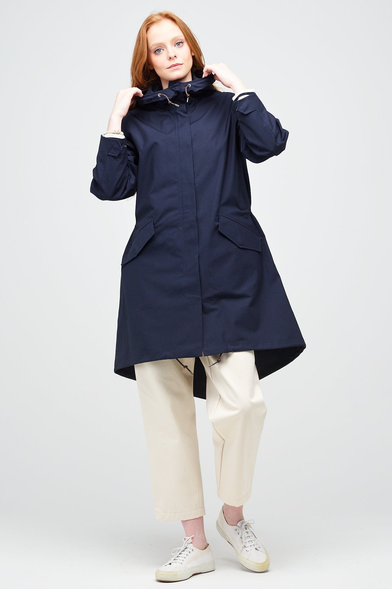A young, white female model with ginger hair wearing a long navy parka fully zipped up. The parka is styled with ecru jeans and white trainers..