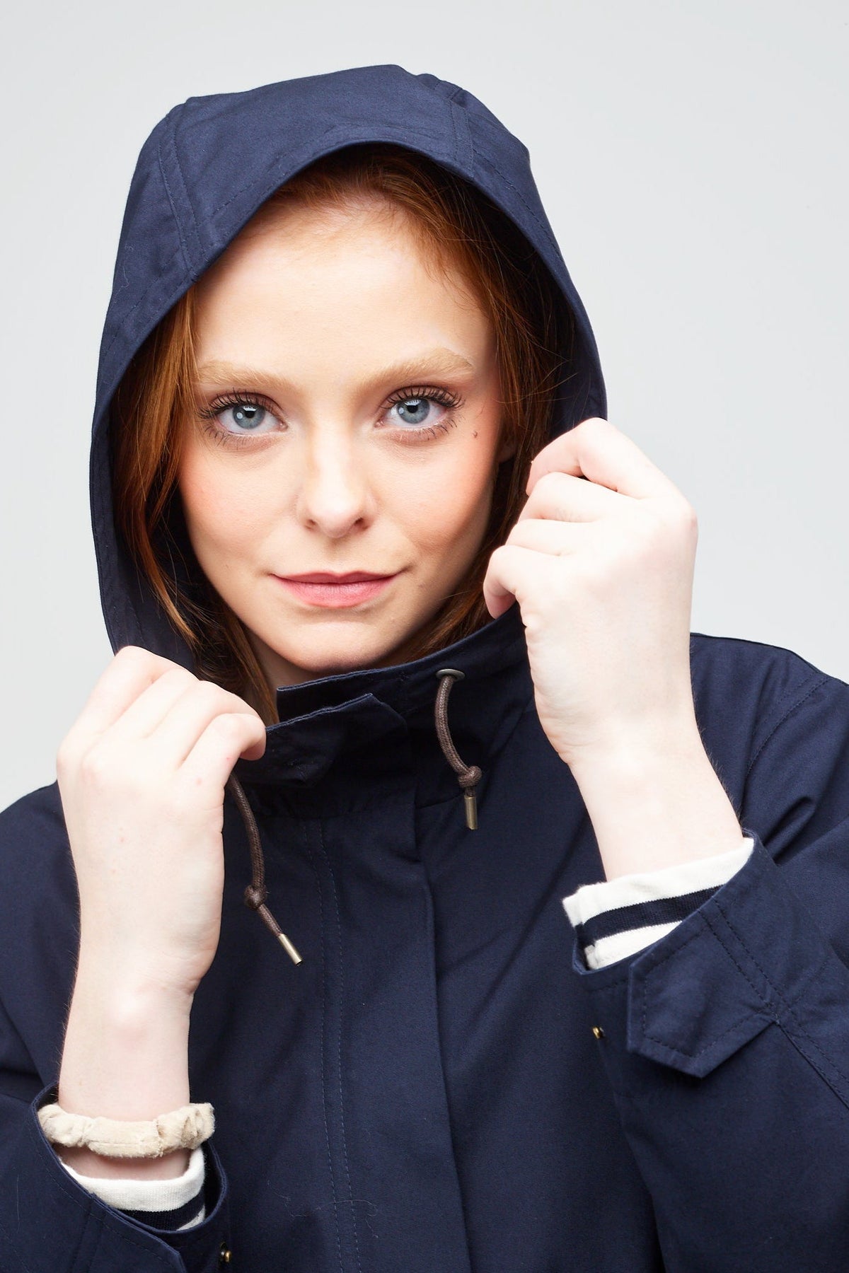 
            shoulders up image of a young, white model with ginger hear wearing a navy long parka with the hood up