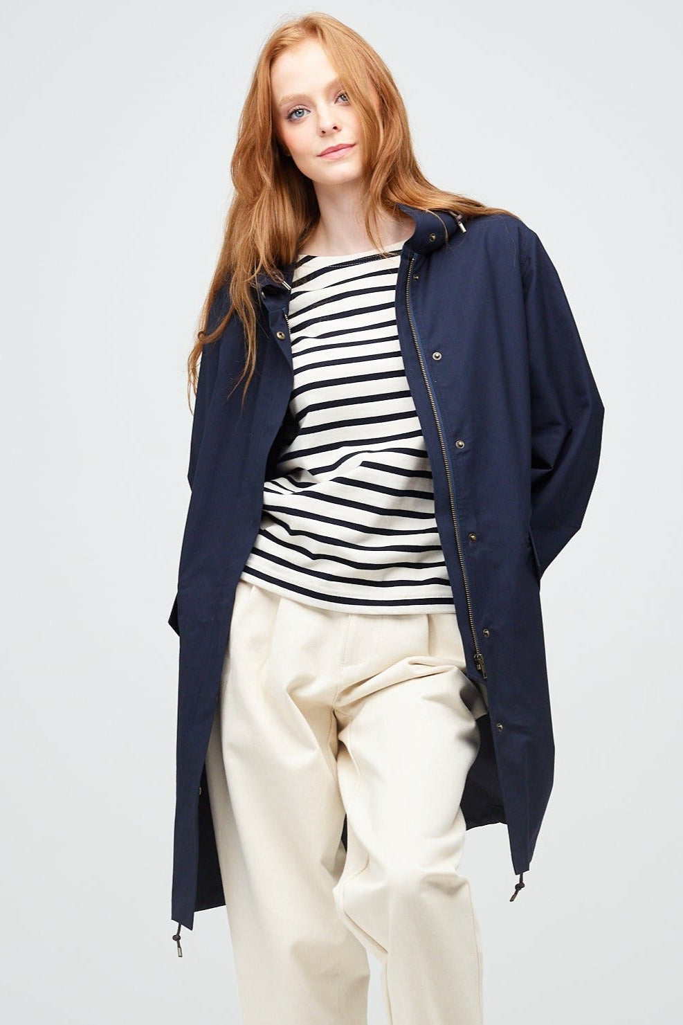 
            A young, white female model with ginger hair wearing a long navy parka unzipped. The parka is styled with a navy striped breton top &amp; ecru jeans.