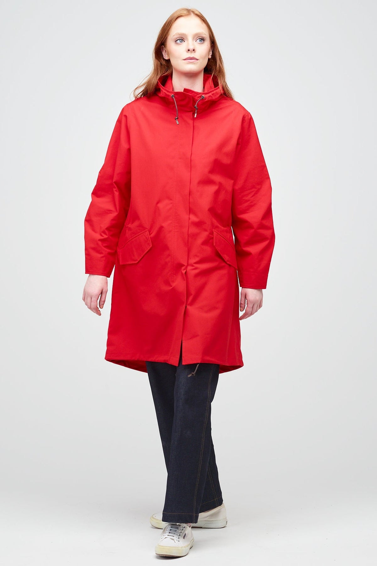 
            A young, white female model with ginger hair wearing long red parka fully zipped up. The parka is styled with indigo jeans and a white trainer.