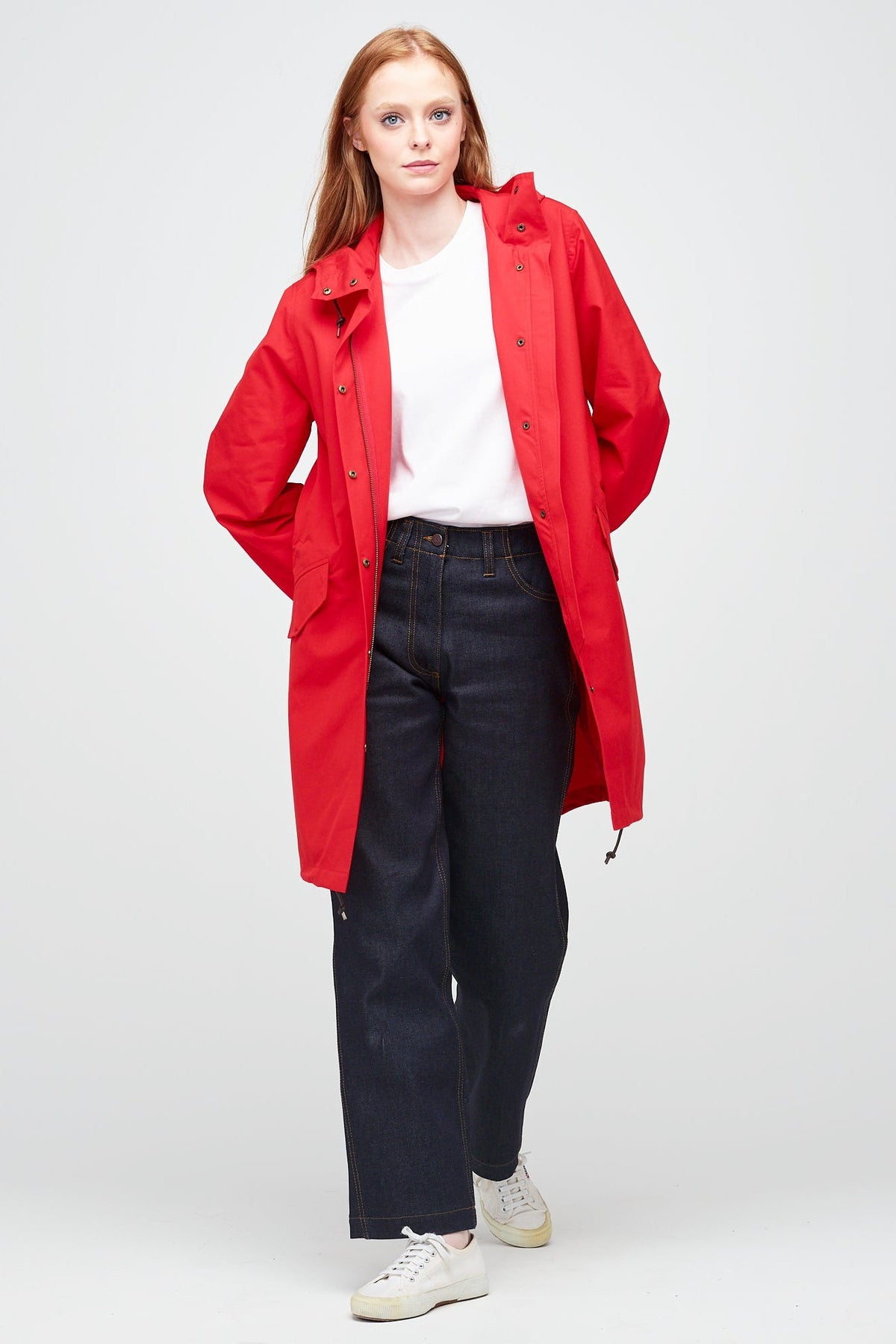 
            A young, white female model with ginger hair wearing an unzipped long red parka. The parka is styled with indigo jeans and a white trainer.