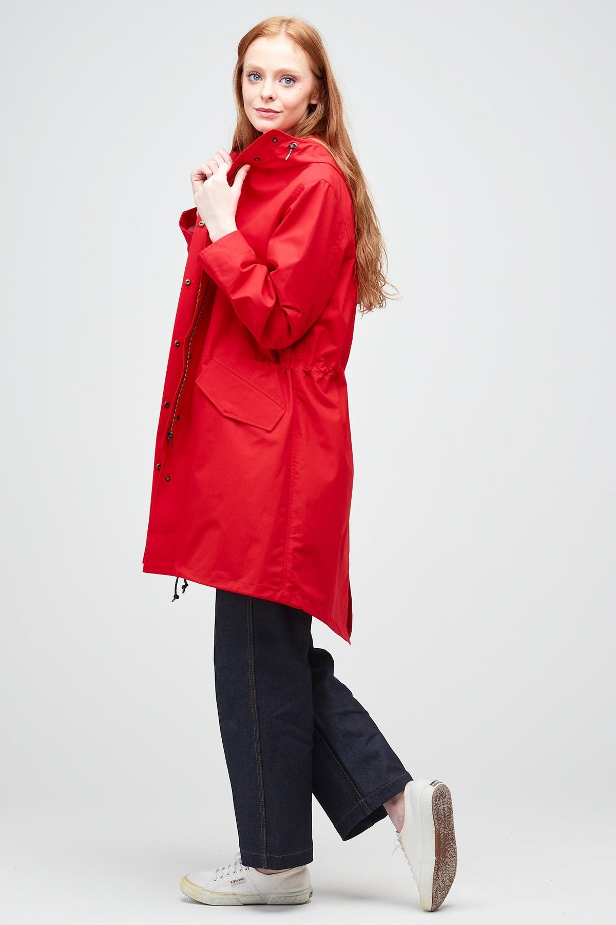 
            Side shot of A young, white female model with ginger hair wearing long red parka unzipped. The parka is styled with indigo jeans and a white trainer.