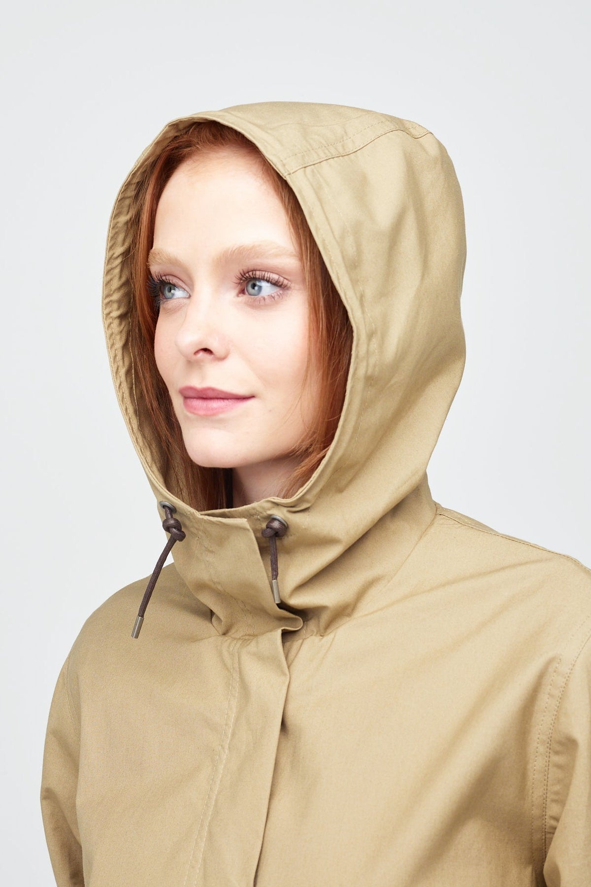 Shoulders up image of a young, white female model with ginger hair with the hood up on a long parka in stone.