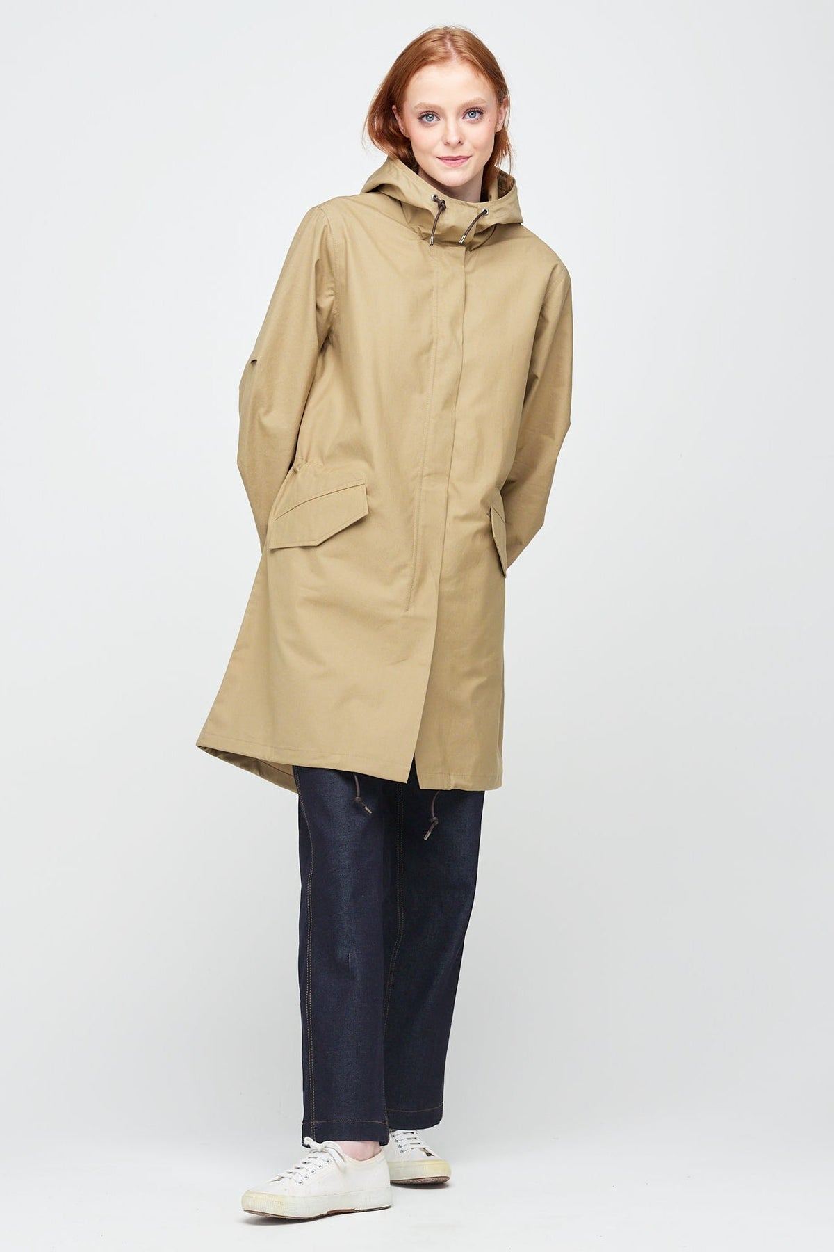 
            A young, white female model with ginger hair wearing long stone coloured parka fully zipped up. The parka is styled with a indigo jeans and a white trainer.