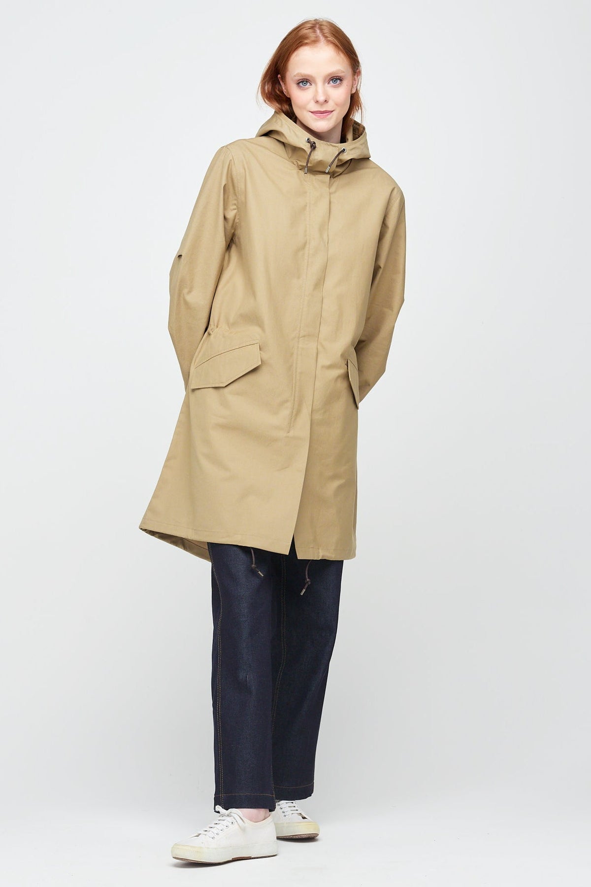 A young, white female model with ginger hair wearing long stone coloured parka fully zipped up. The parka is styled with a indigo jeans and a white trainer.