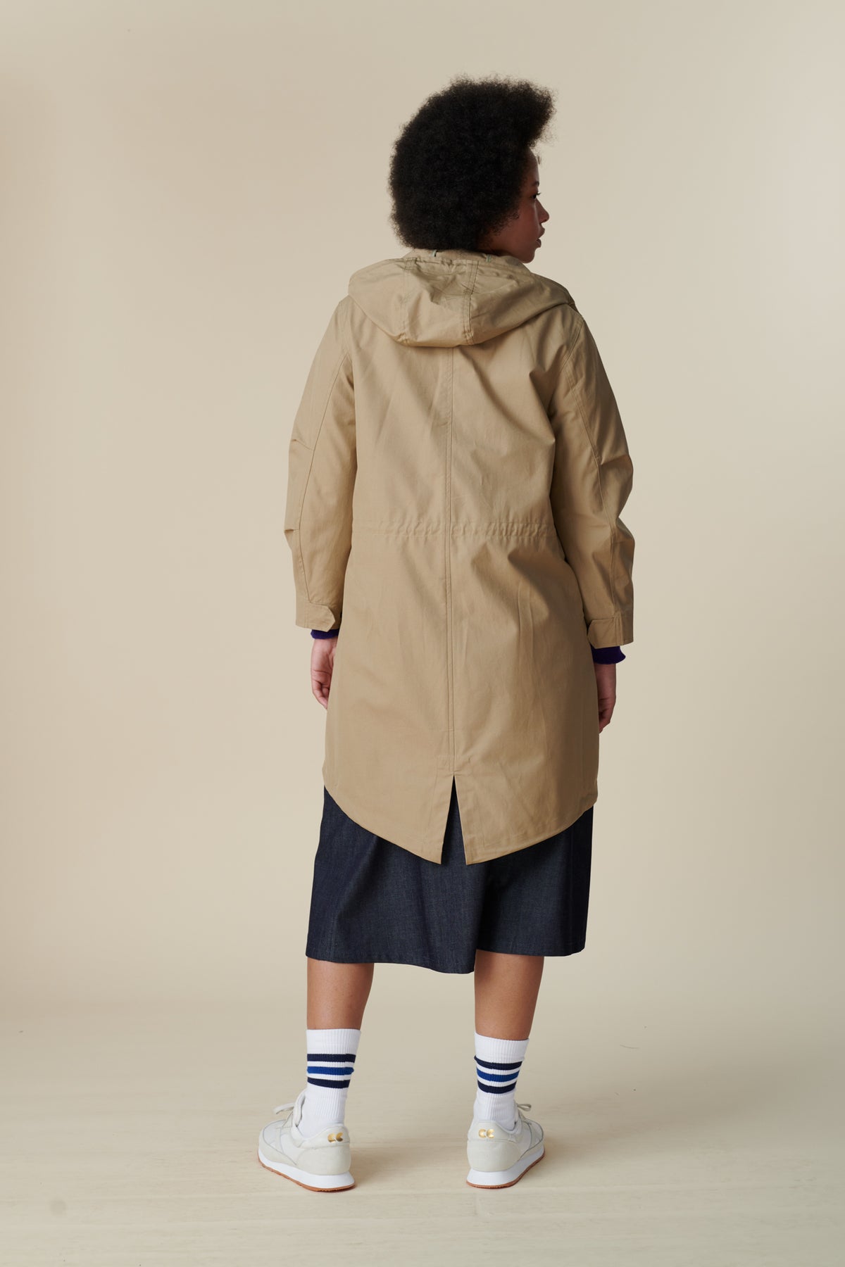 Back shot of black female model with afro wearing long parka in stone over indigo midi skirt